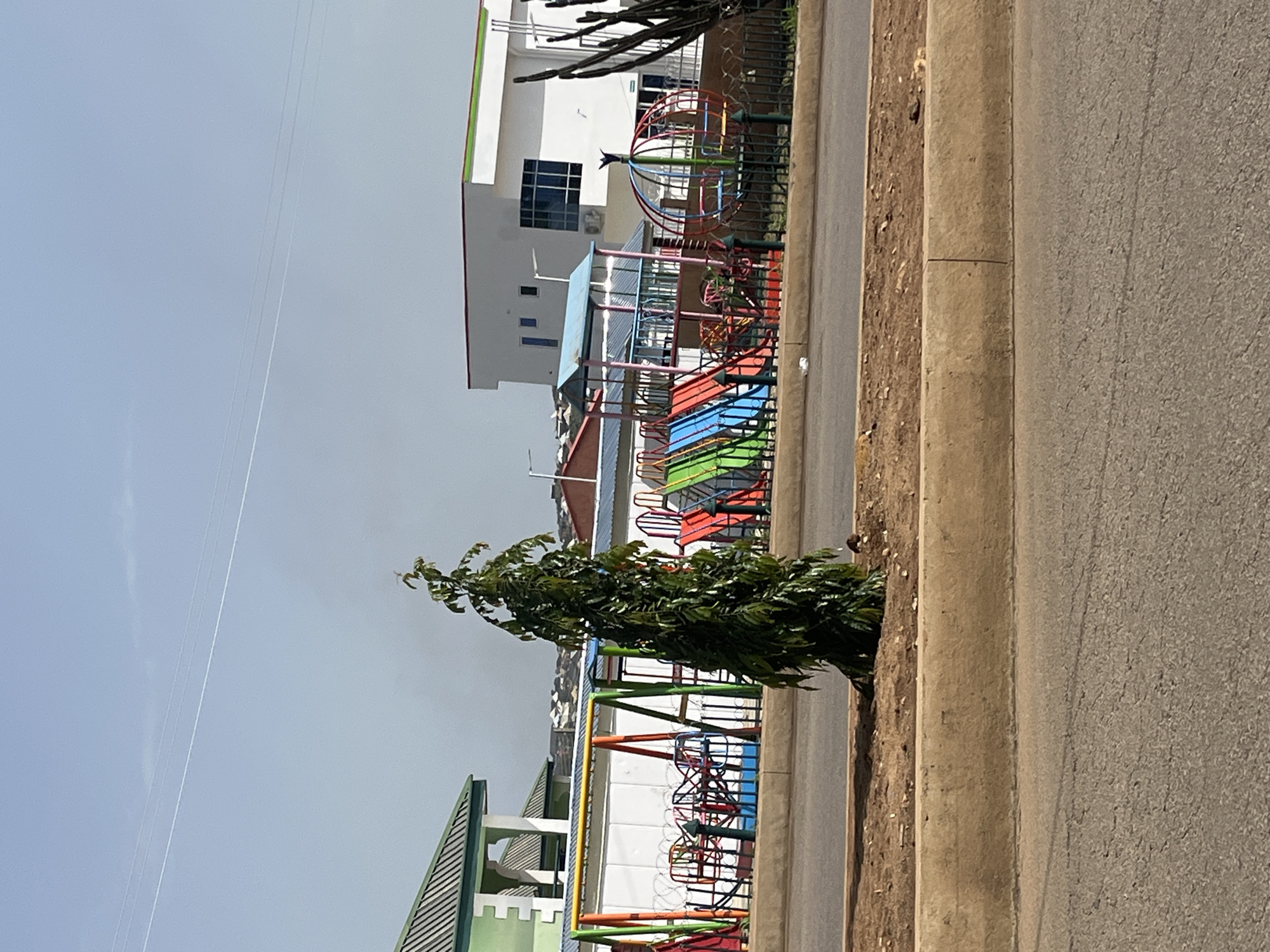 The image depicts a children's playground located in Bukuru, Nigeria. The playground features several colorful pieces of equipment, including slides, swings, and a spherical spinning ride. A tall, slender tree stands prominently in the foreground, casting a shadow on the surrounding dirt and grass. Behind the playground, a white building with a green trim is visible, as are other structures in the background, suggesting an urban or suburban setting. A road runs in front of the playground, separated by a concrete barrier and a strip of dirt. The sky above is a pale blue with some faint clouds. There are no people visible in the image.