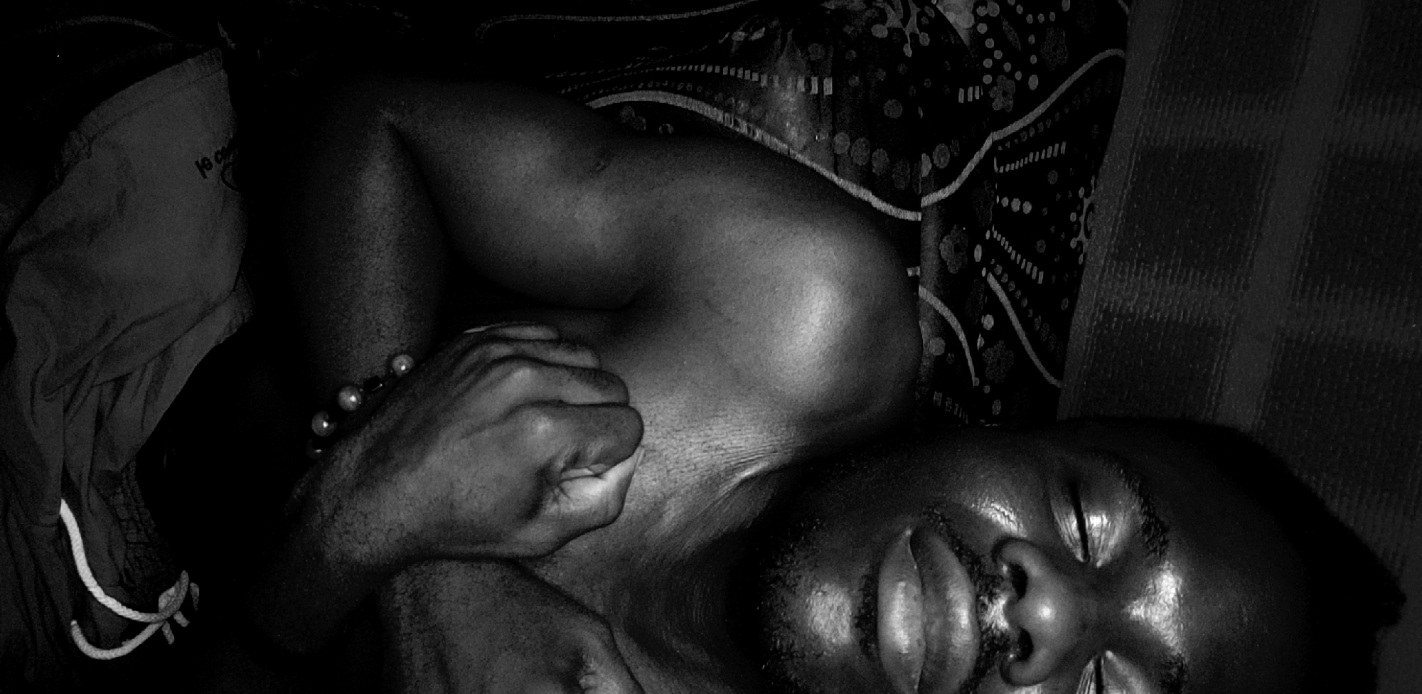 A black and white image depicts a person, likely male, with dark skin, resting or sleeping. The individual is shown from an overhead perspective, lying down with their eyes closed, conveying a sense of tranquility or deep rest. Their upper body is visible, with one arm bent, the hand resting near their chest or neck. A beaded bracelet, featuring light-colored beads, is worn on the visible left wrist or forearm.
The person appears to be on a bed or similar surface, with a dark, subtly patterned fabric providing a background. To the left, a piece of clothing, possibly shorts or trousers, is visible, characterized by a white drawstring and a small label with partially legible text that appears to read "Jeans Co." or similar branding.
The lighting is low and dramatic, creating strong contrasts between highlights on the skin of the face, shoulder, and arm, and deep shadows elsewhere. A distinct sheen is visible on the skin, particularly on the forehead and shoulder, which might suggest warmth or perspiration. The overall composition is intimate and quiet. The scene is set in Narayi, Nigeria.