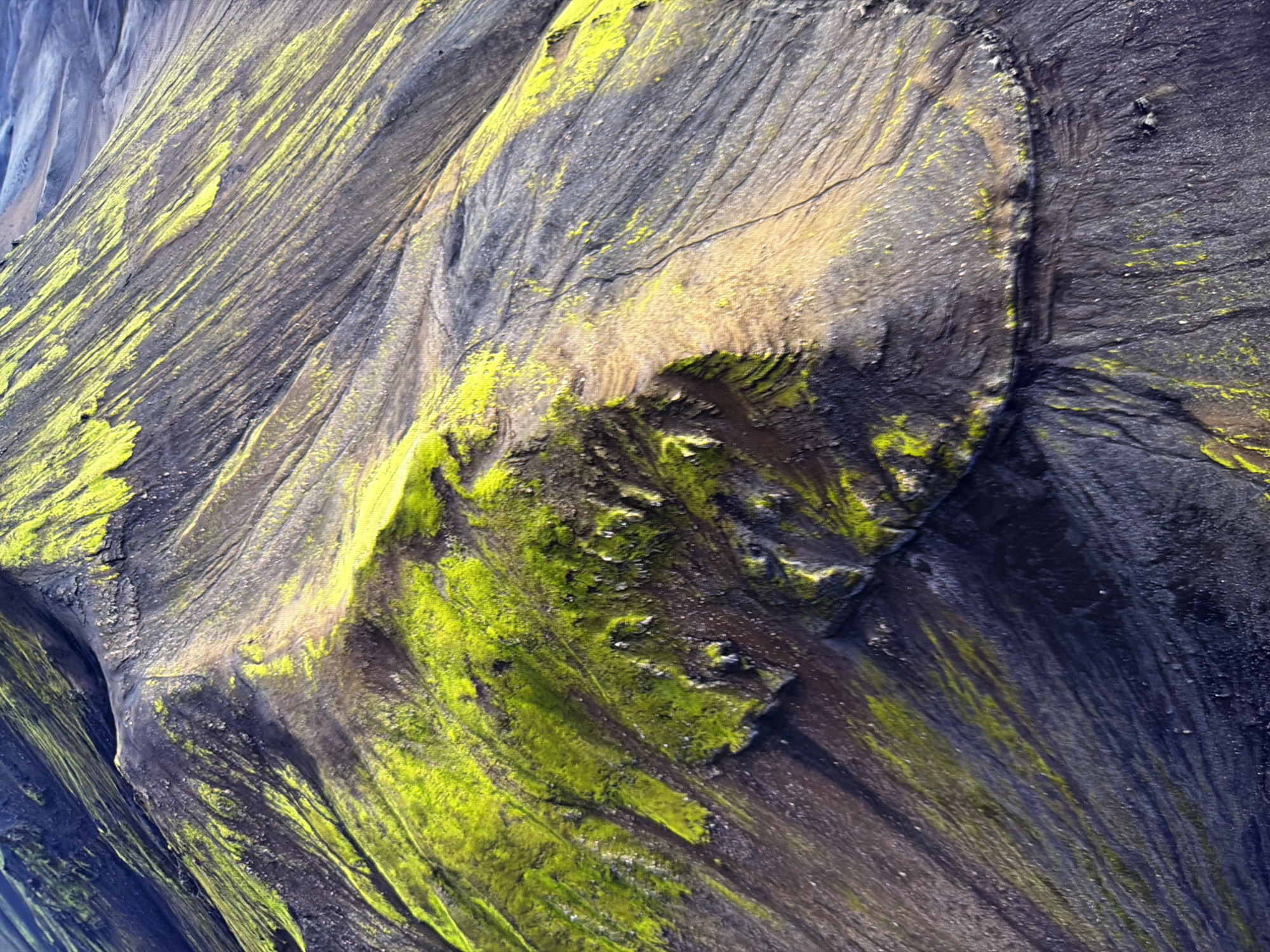 This image is an aerial view of a mountainous landscape covered in dark soil and patches of vibrant green moss. The terrain features steep slopes with visible erosion patterns and streaks of lighter colored earth. The moss grows in irregular clusters and strips, creating a stark contrast against the darker ground. The image does not depict any urban environment, nor does it show any elements related to the city of Kyiv, Ukraine. It is a natural landscape photograph.