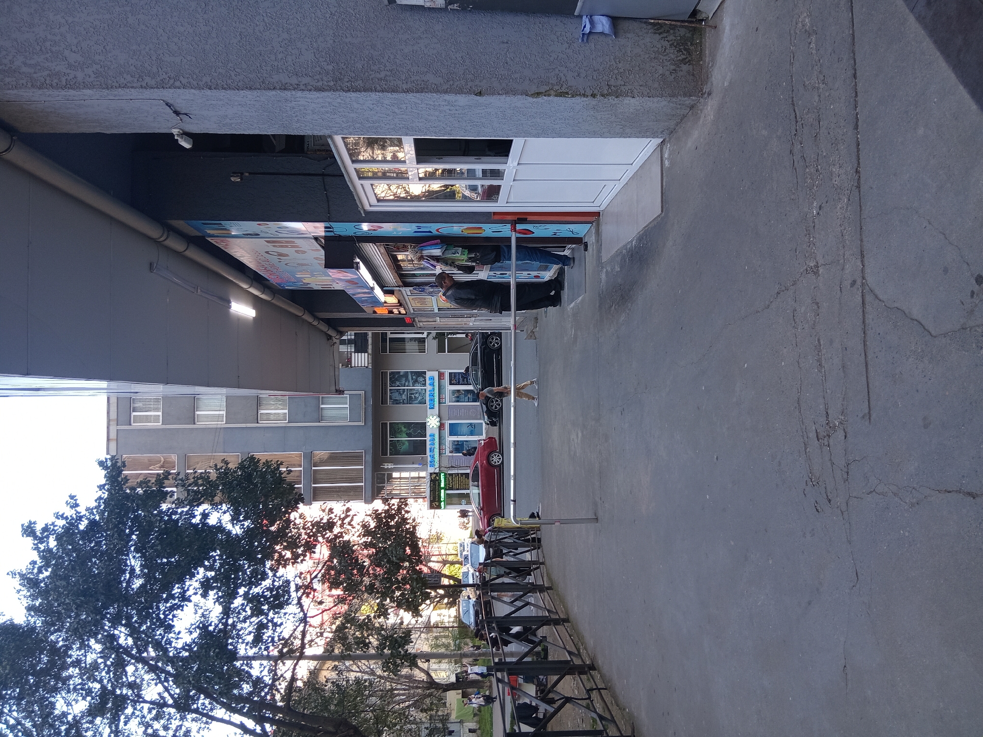The image depicts a pedestrian walkway adjacent to a row of shops in Batumi, Georgia. Two individuals are visible near the shops, one standing and the other possibly seated. A red car is parked along the street. A low black metal fence separates the walkway from a park area with trees. The buildings in the background have a grey facade with windows. The pavement of the walkway is grey and cracked in places.