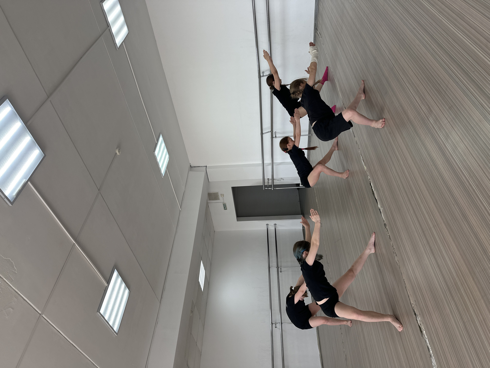 The image shows a dance studio in Votkinsk, Russia. There are four girls, presumably dancers, in the room, practicing at a barre. All are barefoot and wearing black t-shirts and shorts. One has a pink sock on her foot, and another has a bandage on her arm. The studio has gray walls and a light wood-toned floor. The ceiling is equipped with fluorescent lights in square fixtures. There is a metal barre along the wall.