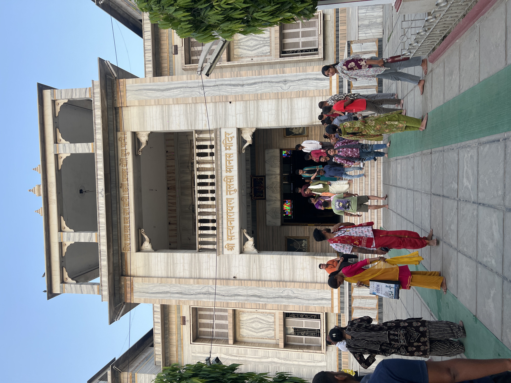 The image shows a group of people lined up in front of a building. The building is made of white marble and has a large entrance. There are two balconies on the left side of the building. There are two white marble sculptures. Above one sculpture is the name written in Hindi of "Sri Satyanarayan Tulsi Manas Mandir" indicating it is a temple in Varanasi, India. There are multiple windows on the front of the building with white shutters. The people in the line are mostly barefoot and are wearing a variety of colorful clothing. There is a green rug rolled out in front of the entrance of the building. The sky is blue in the background.