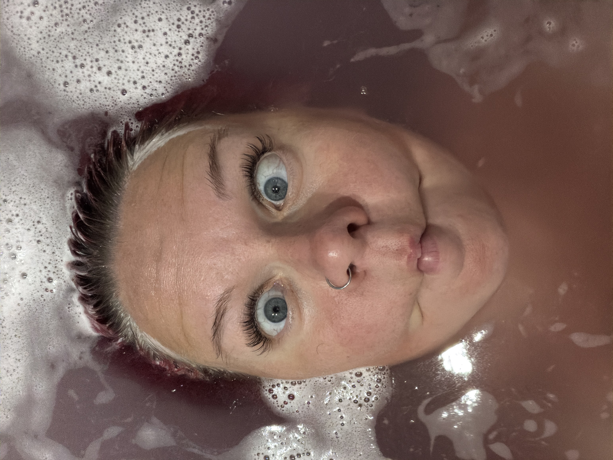 A close-up, high-angle shot shows a person's face emerging from a bath filled with reddish-purple water and white bubbles. The person has light blue eyes, wide with a surprised expression, and is making a fish-like face. Their hair is dark with streaks of bright pink and white, and they are wearing a silver hoop nose ring. The bath water is murky and reflects light, creating a shimmering effect. The scene appears to be a bathroom, likely in a residential setting. There is no indication of the city of Chaikovsky, Russia, in this image.