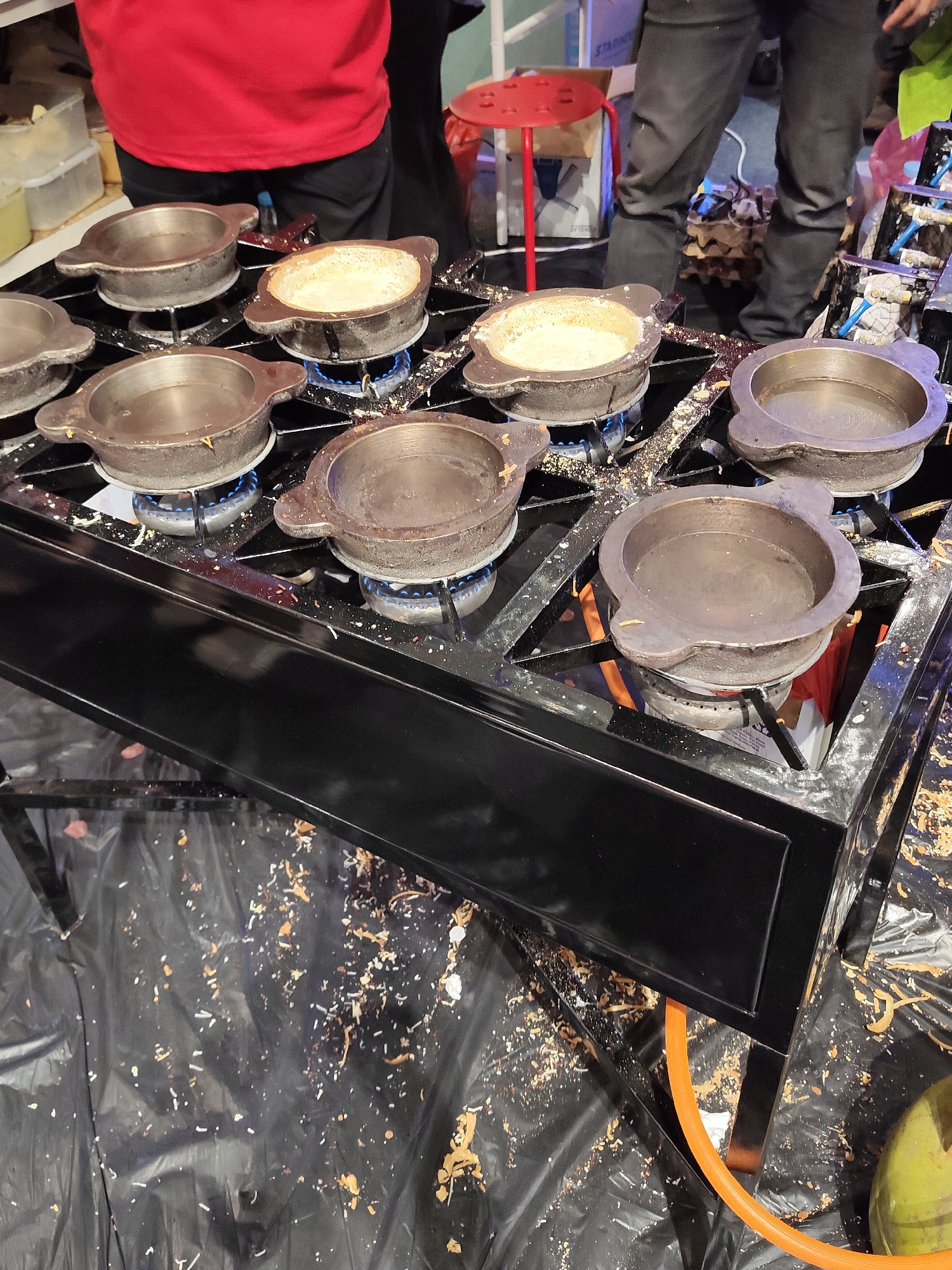 An outdoor cooking station is set up with a black metal stove that has multiple circular heating elements. On the stove, there are several cast-iron molds that appear to be used for cooking pancakes or small cakes. Some of the molds contain batter, while others are empty. A person wearing a red shirt and black pants is visible in the background, suggesting a food stall or a street food vendor. The ground is covered with a black plastic sheet that is littered with food scraps, indicating a busy food preparation area. In the lower right corner, a green coconut is partially visible. The overall scene suggests a food-making operation in a public space.