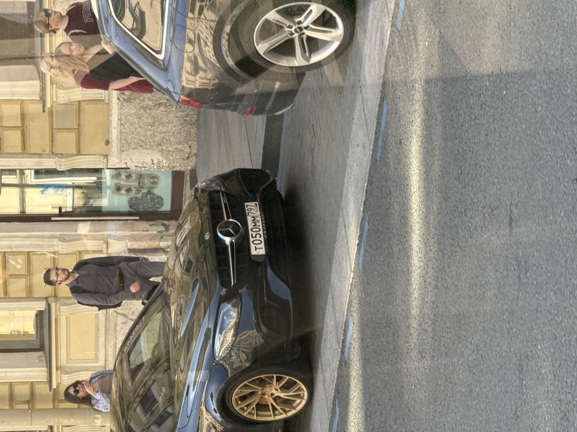 A medium shot captures a street scene in Saint Petersburg, Russia. In the foreground, a black Mercedes-Benz E-Class sedan is parked, its front wheel with a gold rim visible. The license plate reads "T050MM797". To the right, the rear of a dark-colored Audi Q7 is parked, its silver multi-spoke wheel prominent. In the background, several people are standing on the sidewalk. A woman in sunglasses and a striped shirt stands near a building entrance. Beside her, a man wearing glasses, a grey shirt, and carrying a backpack walks away from the camera. Further to the right, three women are seen standing, one wearing a maroon shirt and another a dark jacket. The architecture of the building in the background appears to be historical, with yellow facades and classical detailing. The scene is illuminated by sunlight, casting shadows on the asphalt.
