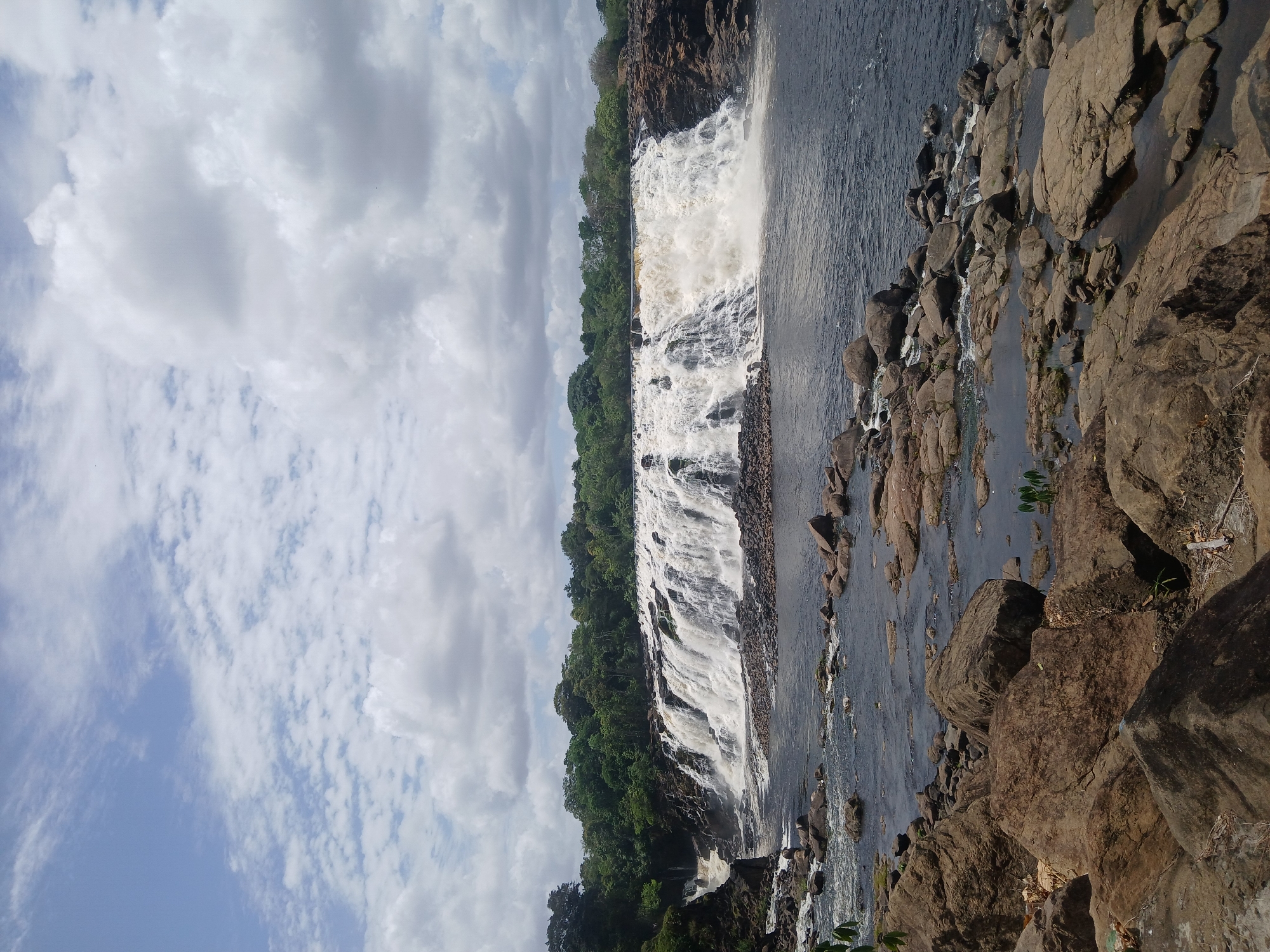 The image captures a wide waterfall flowing into a river. The waterfall is bordered by dense green vegetation on both sides and rocky terrain. The water is churning and foamy as it cascades down the rocks. The foreground consists of large, brown rocks along the riverbank. The sky is filled with scattered clouds.
This image is located in Ciudad Guayana, Venezuela, and is likely Llovizna Falls, within the Llovizna Park.