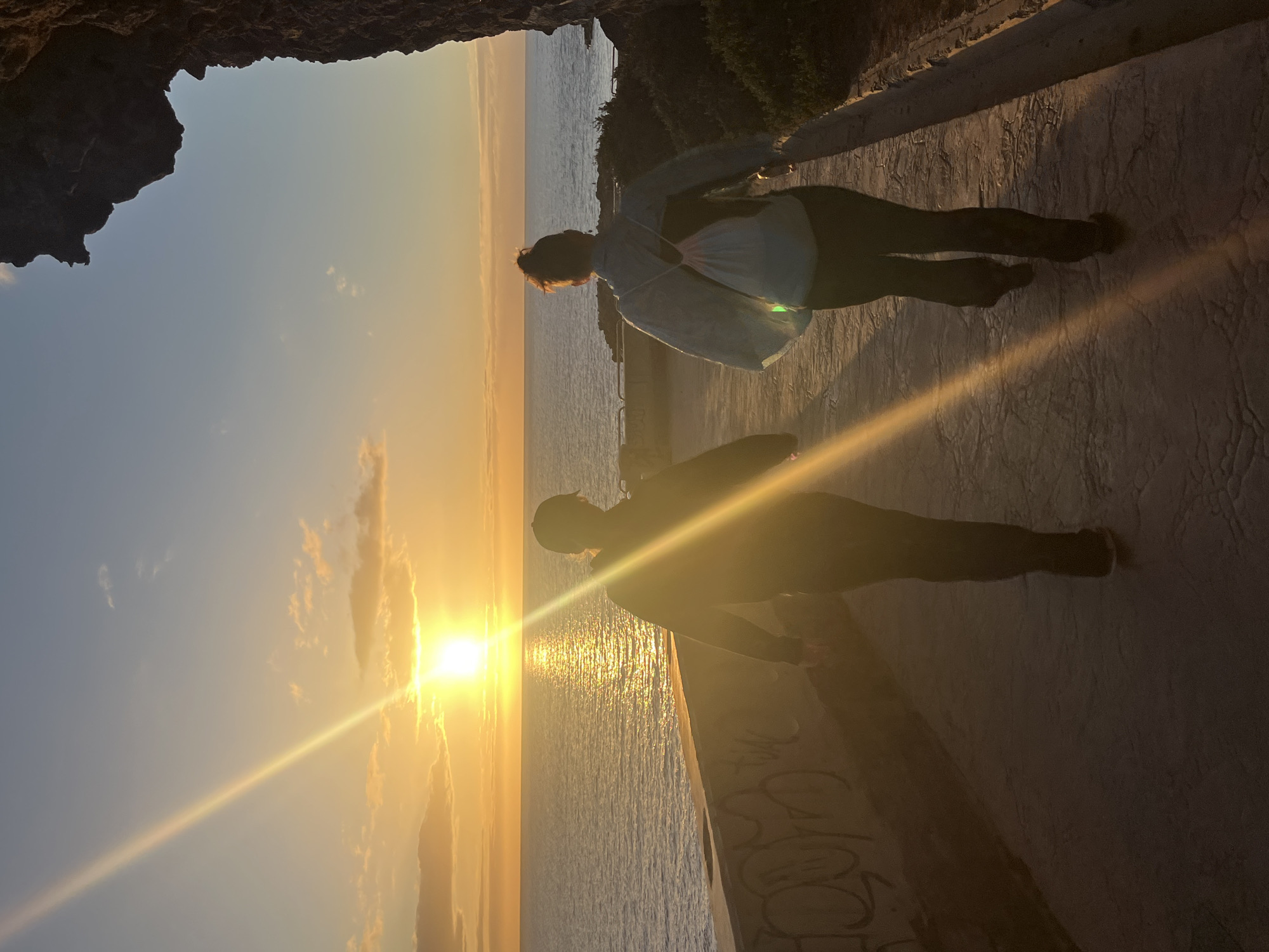 A person in a light-colored shirt and dark pants stands on a textured concrete surface with graffiti along the edge. The sun is setting over the ocean, creating a golden glare and a bright reflection on the water. The silhouette of another person is visible on the concrete surface, along with the person's shadow. The sky is a gradient of blues and yellows. A rocky overhang appears in the upper-left corner of the image. Lush greenery is visible along the upper-right edge. The image appears to have been taken from a high vantage point overlooking the ocean, at sunset. Puerto Rico is located in Gran Canaria, Spain.