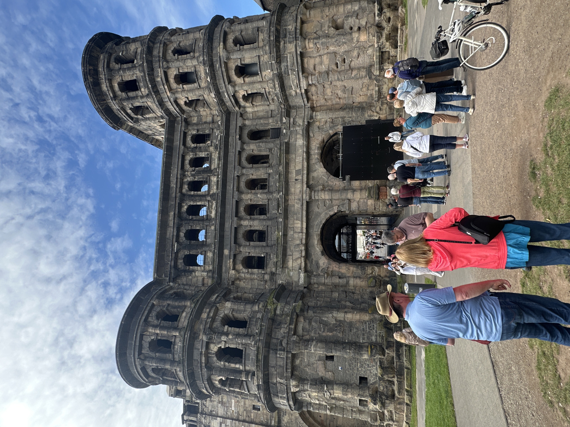 This image depicts the Porta Nigra, a large Roman city gate constructed of dark grey sandstone, located in Trier, Germany. The gate features two imposing, rounded towers with multiple levels of arched openings, flanking a central rectangular structure also with arched windows. At ground level, there are two large archways; the left one is open, revealing people moving through it and a glimpse of a street beyond, while the right archway is blocked by a modern, dark black gate.
Numerous individuals are gathered in front of and around the gate. In the immediate foreground, facing away from the camera, is a man wearing a light blue polo shirt, blue jeans, and a brown cowboy hat. To his right, a woman also faces away, wearing a bright pink jacket over a blue garment, blue jeans, and carrying a black backpack. Several other people, mostly adults dressed in casual attire, are standing in small groups further back, some appearing to be observing the structure or conversing. On the far right, a white bicycle is parked on the unpaved ground. The ground surface in the foreground is a mix of dirt and gravel, with a patch of green grass visible on the left. The sky overhead is partly cloudy with significant patches of blue.