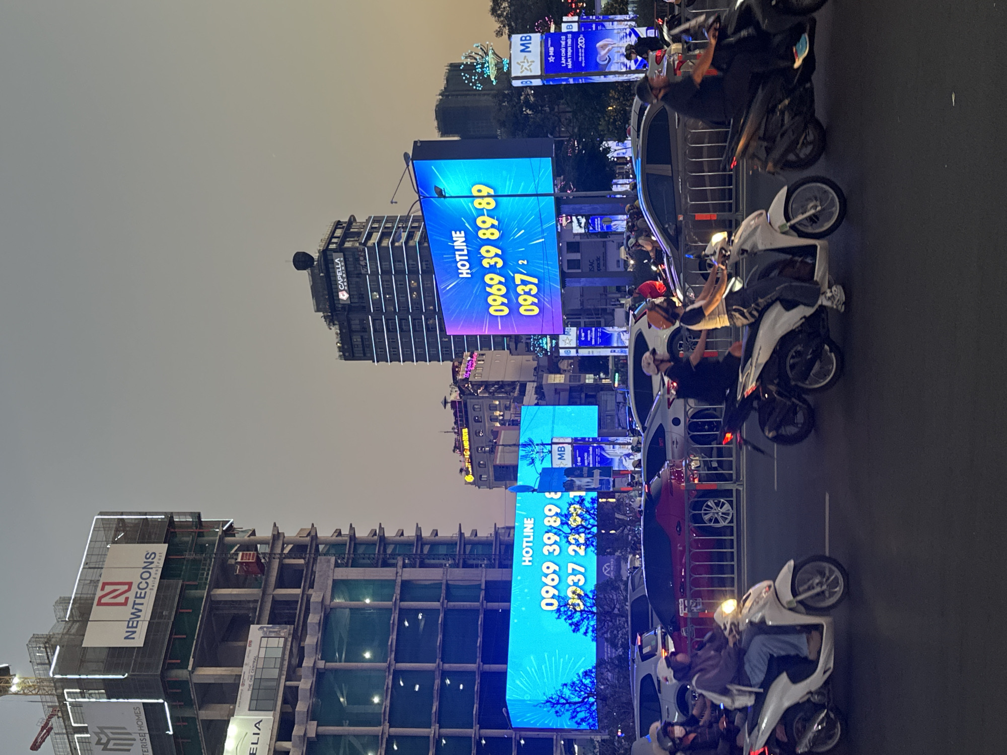 This image captures an urban street scene at dusk or early night in District 1, Ho Chi Minh City, Vietnam. The sky transitions from light orange near the horizon to dark grey overhead, illuminated by numerous artificial lights from buildings, billboards, and vehicles.
The foreground and midground feature a multi-lane road with dense traffic. Approximately ten individuals on motorcycles and scooters are visible, most wearing helmets, and some with face masks. A white scooter with an illuminated headlight is prominent in the immediate foreground, ridden by an individual in a light-colored top and blue jeans. Other scooters are white, black, and dark-colored. Several cars, including a red SUV and silver sedans, are present in various lanes, appearing to be in motion or waiting.
On the left side of the image, a multi-story building under construction, primarily concrete framework and scaffolding, dominates the skyline. It bears large banners displaying "NEWTECONS" (with an 'N' logo and "Build on trust"), "ENTERPRISE HOMES," "CELIA," and "THE ONE CENTRAL," identifying it as the "The One Central" project. To the right, a modern, dark-colored high-rise building with "CAPELLA HOLDING" signage at its peak is illuminated with horizontal light strips.
Two large, luminous digital billboards are prominent, displaying a blue and purple gradient background. Each features "HOTLINE" in white text and two yellow phone numbers: "0969 39 89 89" and "0937 22 99 1" (partially visible on the right as "0937 2"). Smaller vertical light poles with "MB" logos and Vietnamese text are also present. In the background, additional city buildings are visible, some with illuminated signs, including one that reads "GRAND HOTEL." The scene indicates an active urban environment with significant commercial development.