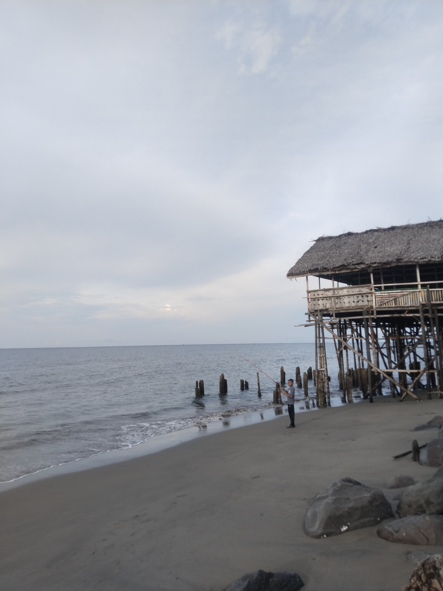 A young man stands on a sandy beach, fishing with a red fishing rod in the shallow water. The sky above is overcast with a pale, hazy light. To the right of the frame, a stilted structure with a thatched roof extends out over the water, supported by numerous wooden posts. Several wooden pilings are visible in the sea near the man and the structure. Large rocks are scattered on the beach in the foreground. The scene appears to be located on a coastal area in or near the city of Lhokseumawe, Indonesia.