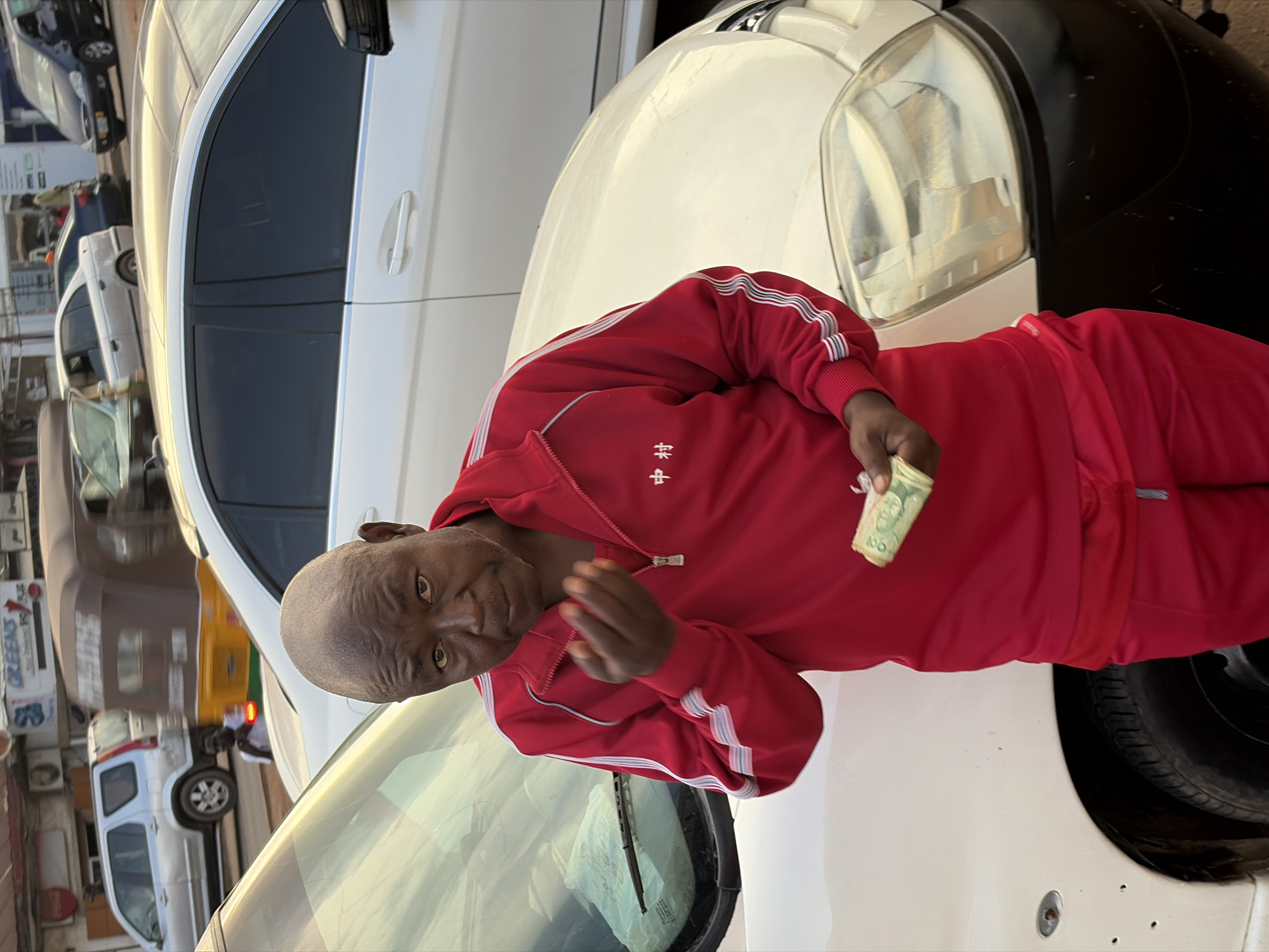 A man in a red tracksuit stands in front of a white car, holding a rolled-up bill in his right hand and gesturing with his left. The man, appearing to be middle-aged with a shaved head and dark skin, has a focused expression as he looks towards the camera. His tracksuit features white and grey stripes on the sleeves and a small embroidered symbol on the chest. The car is a modern sedan with tinted windows and chrome trim. In the background, other vehicles, including a silver SUV and a colorful auto-rickshaw, are visible, suggesting an urban environment. The setting appears to be a street in Jos, Nigeria, likely a bustling commercial area with shops and traffic. The overall lighting suggests it is daytime, possibly late afternoon due to the warm tones. The image is taken from a medium shot, capturing the man from the chest up, with the car's hood and windshield in the foreground.