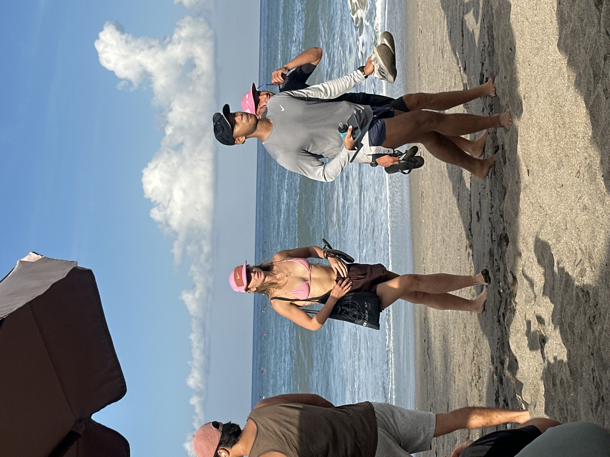 In North Kuta, Indonesia, a group of four individuals are walking on a sandy beach. The foreground is dominated by sand with a shadow cast from an unseen umbrella to the left. In the middle ground, a woman wearing a pink bikini top, brown shorts, and a pink cap walks towards the right, carrying a black tote bag and a pair of flip-flops. To her left, a person wearing a brown tank top and grey shorts faces away from the camera. To the right of the woman, two men are walking. The man in front wears a grey long-sleeved shirt, dark shorts, a black baseball cap, and carries a water bottle and a phone in his hand, along with a pair of flip-flops. Behind him, another man wearing a black shirt and a pink cap is partially visible. The background shows the ocean with gentle waves and a line of surfers in the distance, under a bright blue sky with scattered white clouds.