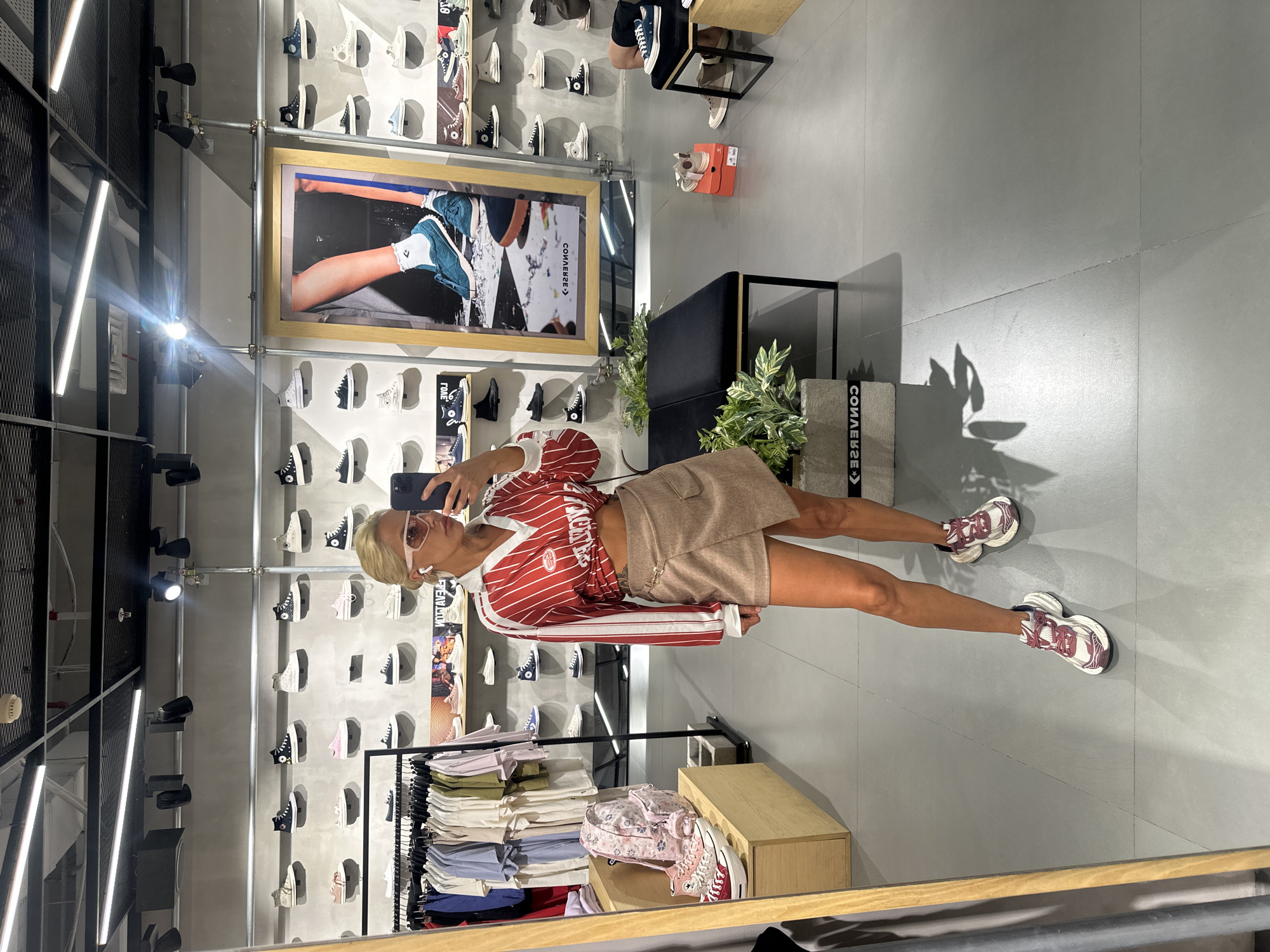 A woman stands in the Converse store at Beachwalk Shopping Center in Kuta, Indonesia, using her phone to take a picture of herself in a full-length mirror. She is wearing a red and white striped shirt, a short khaki skirt, white sunglasses, and chunky maroon and white sneakers. Behind her is a wall displaying various Converse shoes and a framed promotional image featuring feet in Converse sneakers. To her right is a rack of clothing and a low wooden shelf. The floor is light gray tile. Fluorescent lights hang from the ceiling.