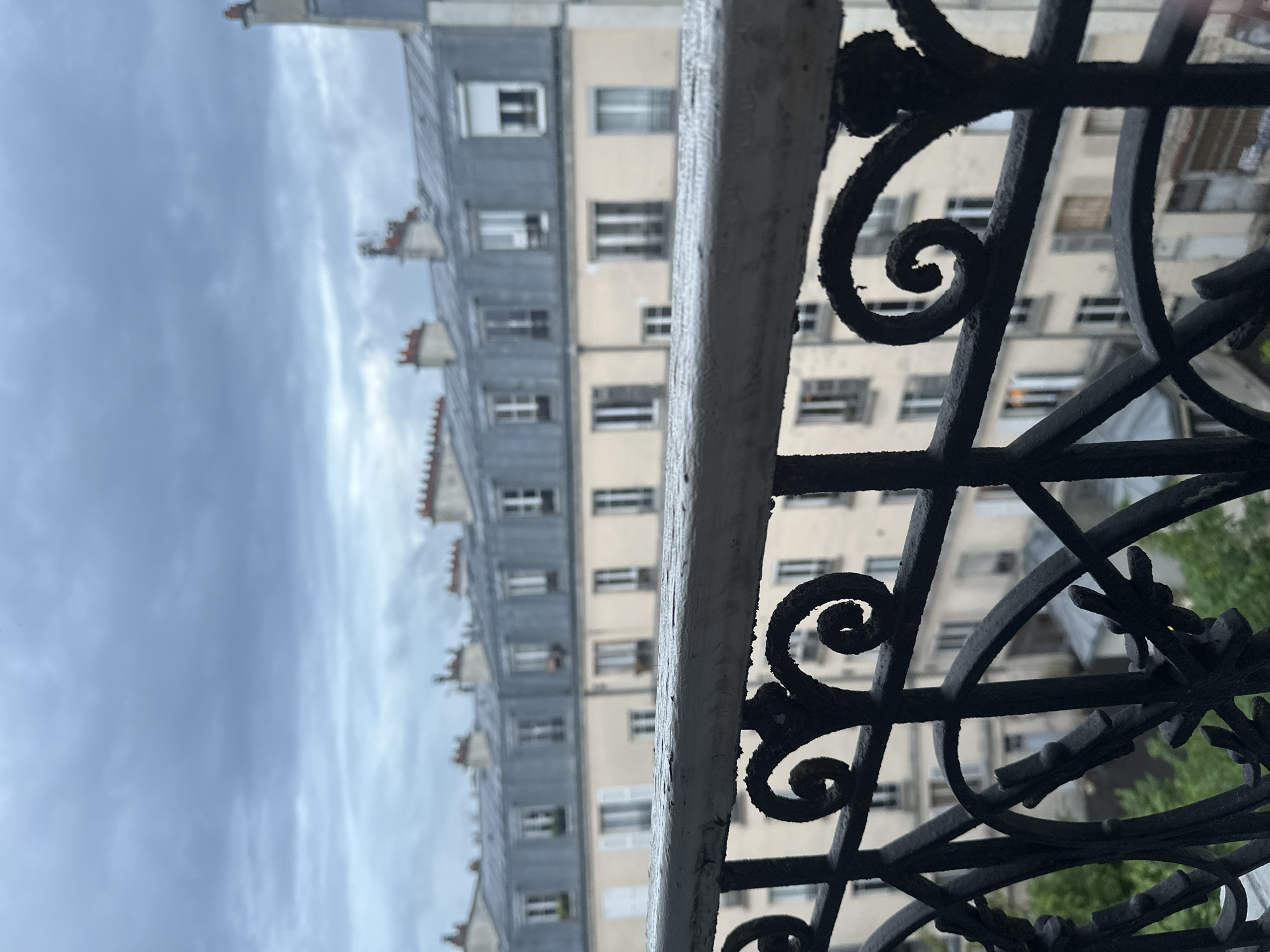 A medium shot captures a view from a balcony in Saint-Ouen-sur-Seine, France. The foreground is dominated by an ornate black wrought iron railing, its design featuring prominent swirls and decorative elements. A white, rectangular support beam runs horizontally across the frame, slightly obscuring the view beyond.
In the background, a Parisian building is visible, characterized by its light beige facade and rows of windows. Above the building, the sky is overcast with gray clouds. The overall perspective is slightly tilted to the left.