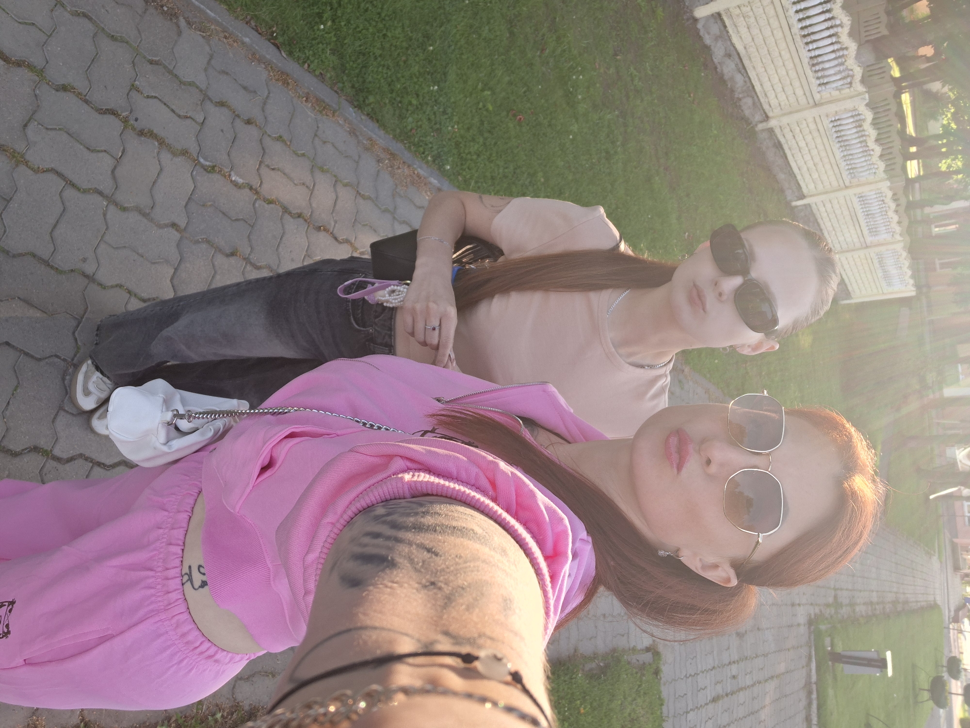 In Myadzyel, Belarus, two women are taking a selfie outdoors. The woman in the foreground, with red hair and sunglasses, is wearing a pink tracksuit and holding a small white purse. The woman behind her, with long brown hair and sunglasses, is wearing a beige t-shirt and dark jeans, and is carrying a black purse. They are standing on a paved walkway next to a grassy area and a light-colored fence. Sunlight is streaming in from the upper left, creating a lens flare effect.
