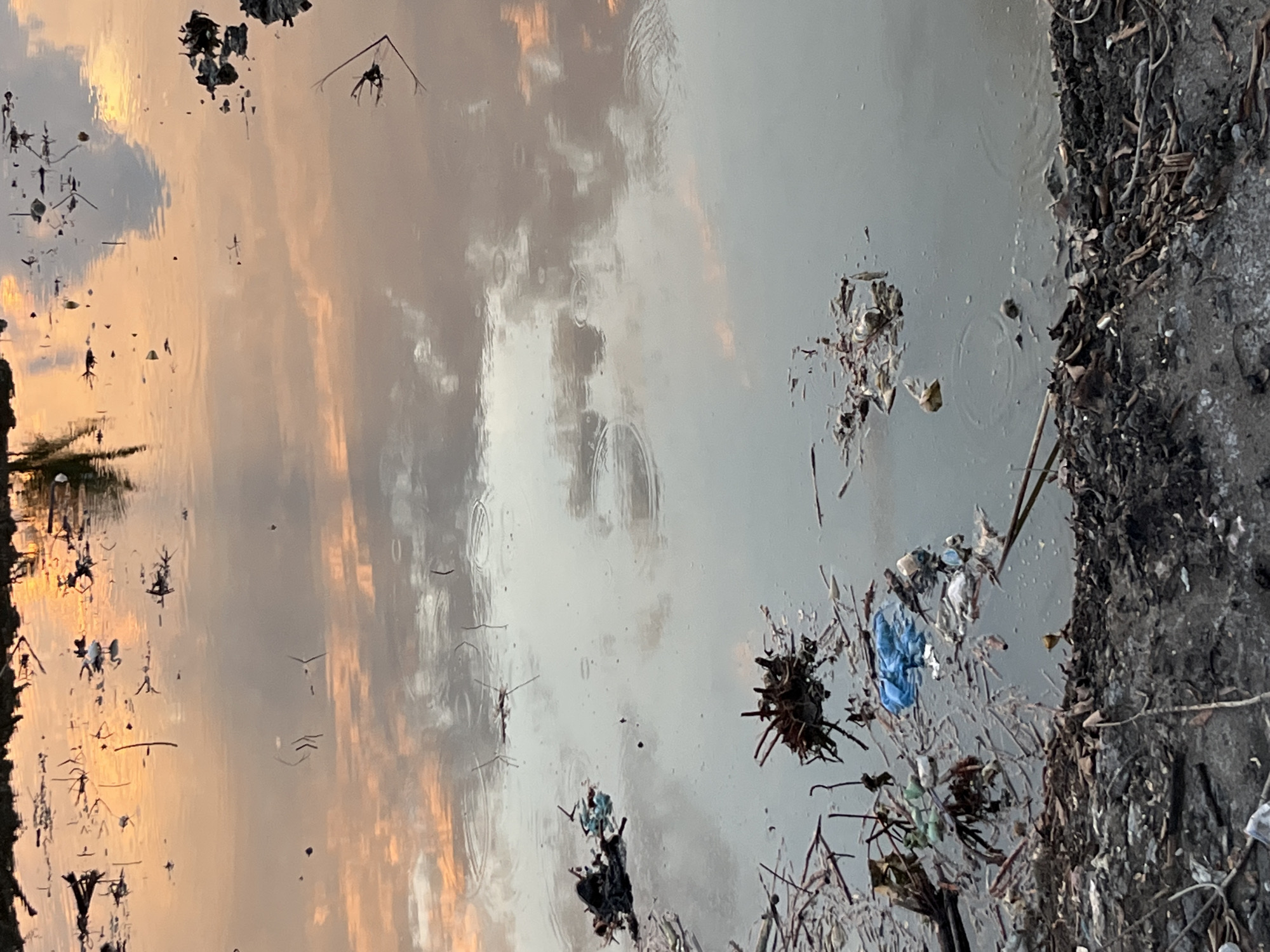 The image shows a body of water at dusk, reflecting the sky's colors of orange and pink. The water is heavily polluted with trash, including plastic bags, debris, and organic matter, floating on the surface and along the bank. The bank itself is covered in a mixture of mud, leaves, and more discarded items. The scene suggests a neglected area, possibly a canal or a low-lying drainage area within the city of Lhokseumawe, Indonesia, likely near a residential or industrial zone where waste management is poor. There are no people visible in the image.