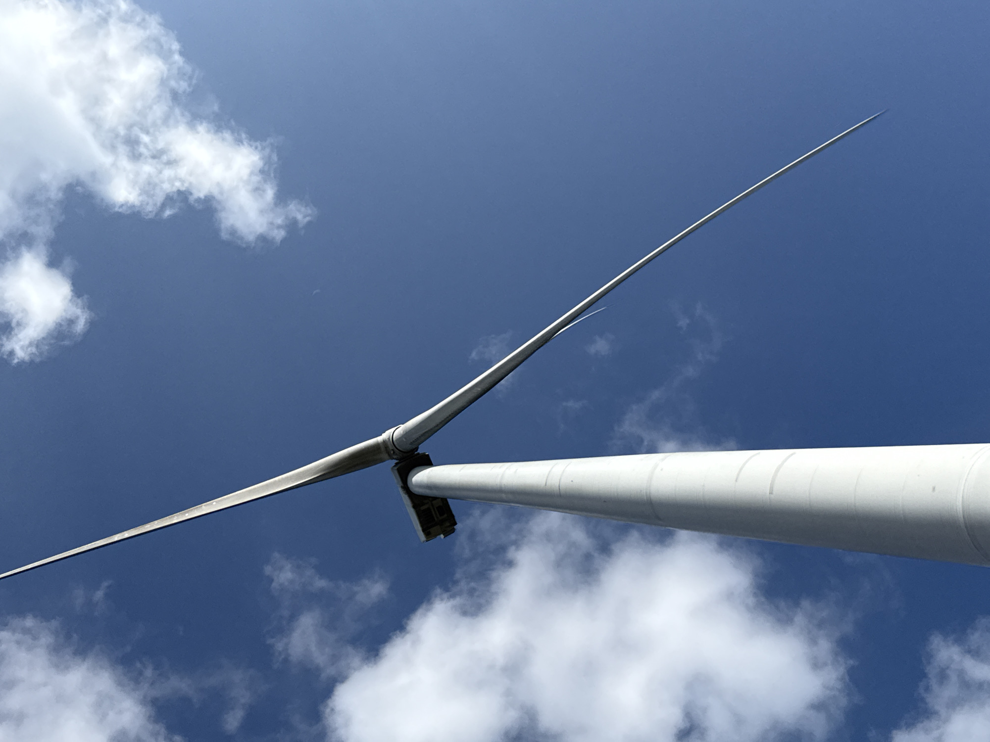 A low-angle, full-frame shot captures a white wind turbine against a clear blue sky with scattered white clouds. The turbine's tall, cylindrical tower dominates the left side of the frame, tapering slightly as it ascends. At the top of the tower, a nacelle is visible, from which two long, slender blades extend diagonally across the sky. One blade points upwards and to the right, appearing to thin to a sharp point in the distance, while the other blade is angled downwards and to the left, disappearing out of the frame. The image appears to be taken from ground level, looking directly up at the structure. The wind turbine is located in Trường Thọ, Vietnam, suggesting an outdoor, possibly industrial or agricultural setting.