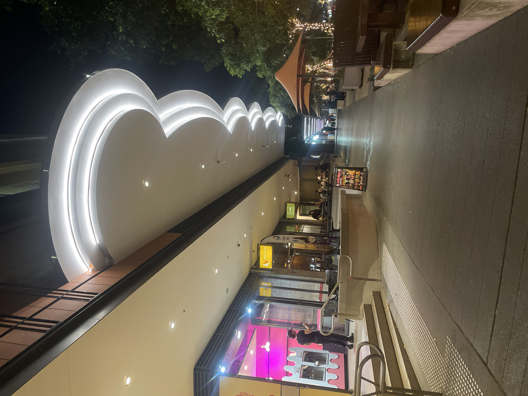 The image depicts a nighttime view of a pedestrian walkway in Brisbane, Australia. The walkway is lined with shops on the left and outdoor seating areas on the right. A distinctive architectural feature is a wave-like canopy with integrated lighting extending over the walkway. On the left, a person in a black uniform is standing in front of a brightly lit shop with pink interior lighting. To the right, there are several outdoor dining tables and chairs, some under orange umbrellas, with decorative string lights illuminating the trees and surrounding area. People are seated at some of the tables. The walkway is paved and has tactile paving indicators near the steps leading up to the shops. A sign with colorful images and text stands near the walkway. The overall impression is of a lively, well-lit urban entertainment precinct at night.