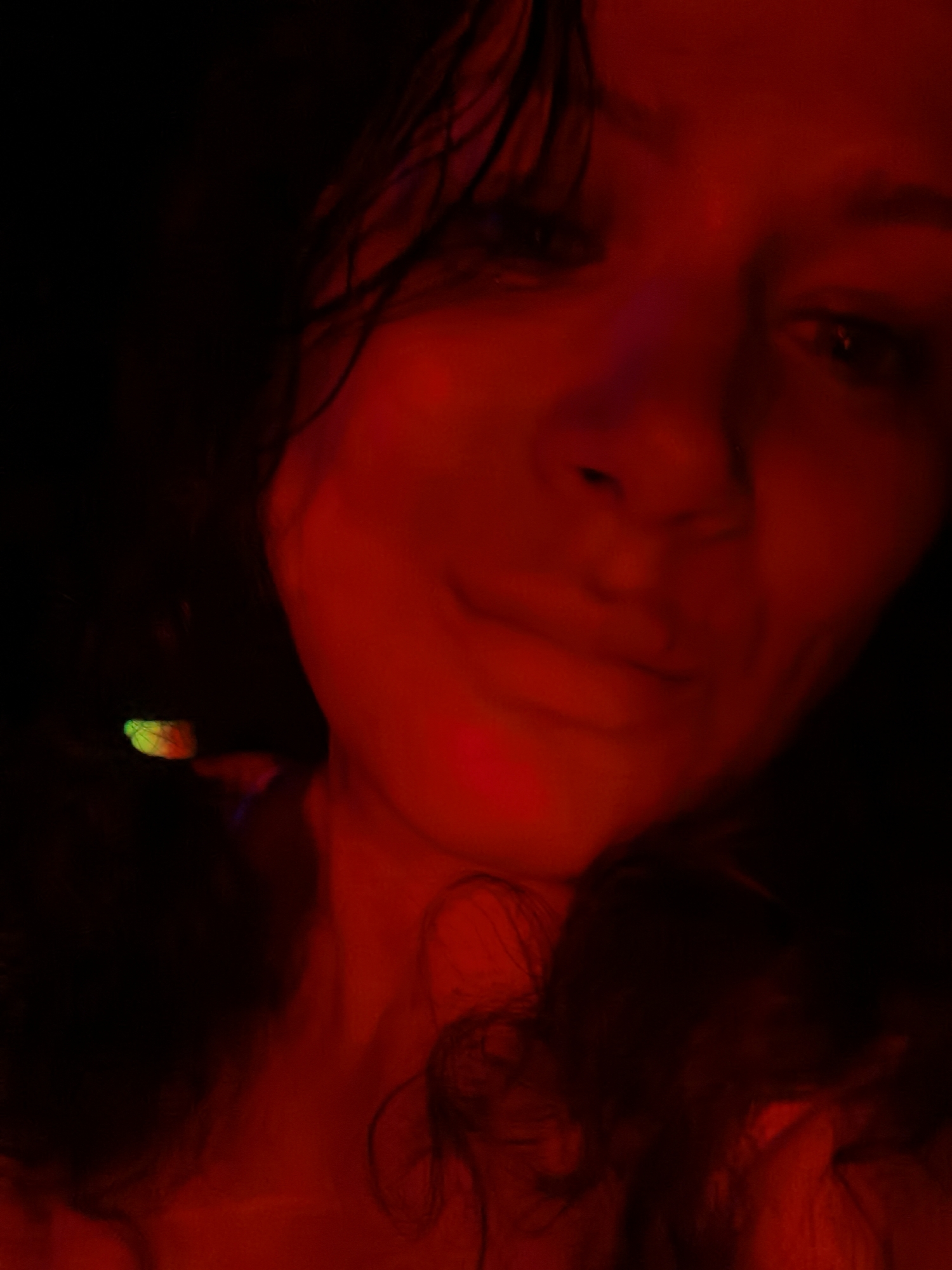 The image is a close-up selfie of a person with medium-length, dark hair. The scene is predominantly illuminated by red light, obscuring details. The person's face is visible, and they are looking slightly downwards with a subtle smile. A small area with green and orange coloration is present near the left side of the image, likely indicating a light source or object. The background is dark and indistinct. The location is not discernible from the image.