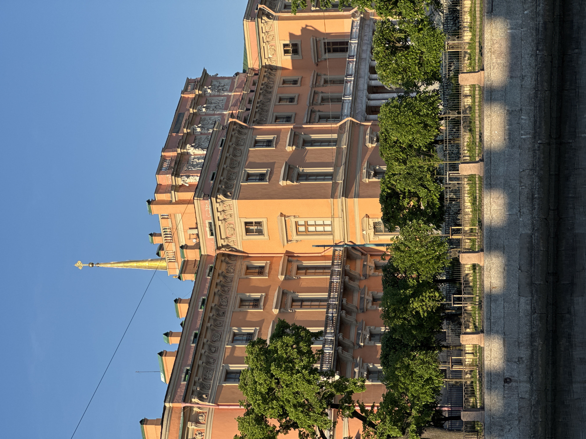This media file features a grand, multi-story building with a distinctive orange-pink facade under a clear, bright blue sky. The architectural style is classical, adorned with intricate details such as decorative window surrounds, cornices, pilasters, and what appear to be sculptural reliefs along the upper stories. A prominent golden spire, capped with a finial, rises from the left side of the building, suggesting a significant landmark or palace.
Green, leafy trees are visible in the foreground and along the base of the building, partially obscuring its lower levels. In the bottom right of the image, a metal fence with stone posts runs along a paved area, which appears to be a canal or river embankment. Long, distinct shadows are cast across the paved surface, indicating a low sun angle, suggesting either late afternoon or early morning. The sunlight warmly illuminates the building, enhancing its colors and highlighting its architectural features.
The scene depicts an urban setting, characterized by impressive historical architecture and waterways, consistent with Saint Petersburg, Russia. The building's appearance, particularly its color and spire, strongly identifies it as the Mikhailovsky Castle (St. Michael's Castle). No specific activity or event is taking place, and no visible text is present. The overall impression is one of historical grandeur and architectural beauty under clear, sunny conditions.