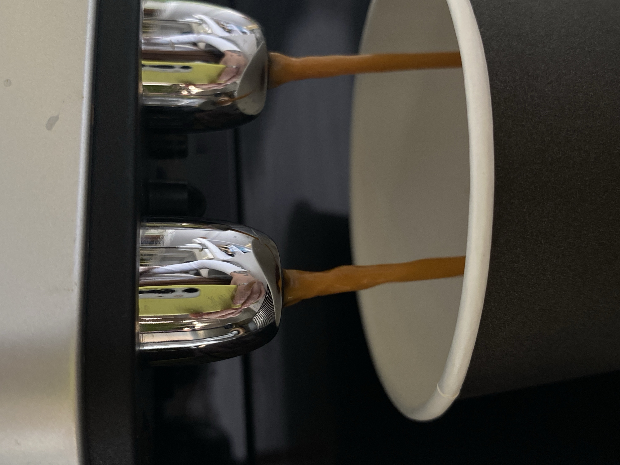 The image shows an espresso machine dispensing coffee into a black and white paper cup. The scene is close-up, focusing on the coffee stream and the portafilter spouts. Reflections in the chrome spouts show a person holding a bright yellow smartphone, suggesting the photo was taken by the user of the machine. The background is dark and out of focus. There is no indication of the city of Votkinsk, Russia, or any other specific location.