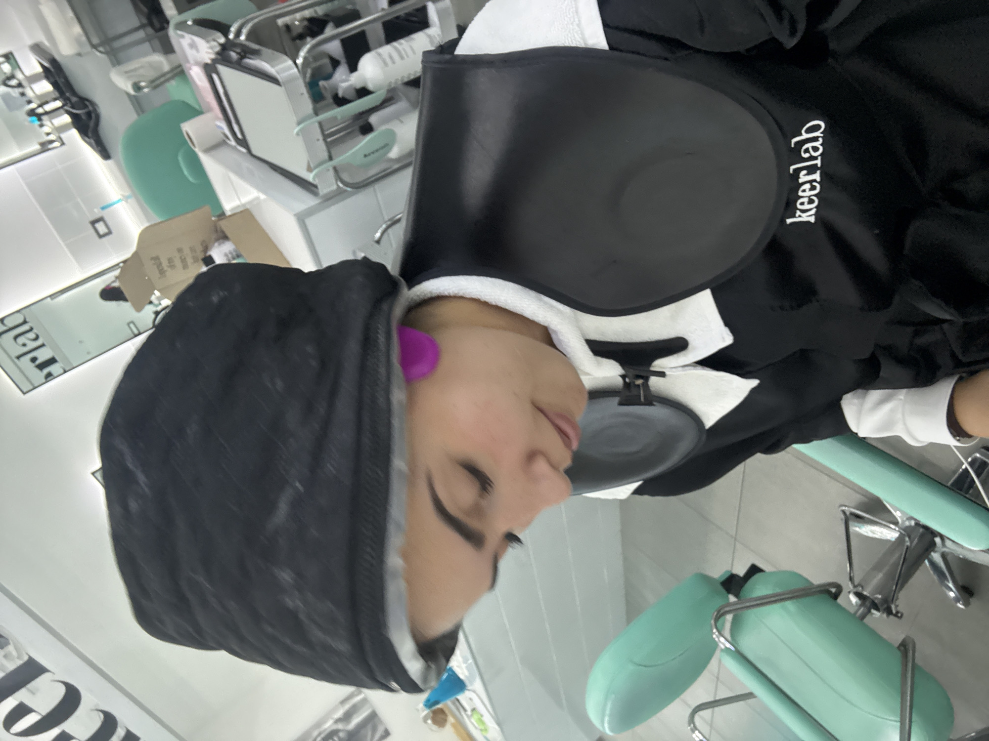 A woman is sitting in a salon chair, wearing a black hair wrap and a black smock with white trim and the word "keerlab" printed on it. She has purple ear protection. The salon has light green chairs and a white counter with salon supplies. The location is a hair salon in San Bernardino Tlaxcalancingo, Mexico.