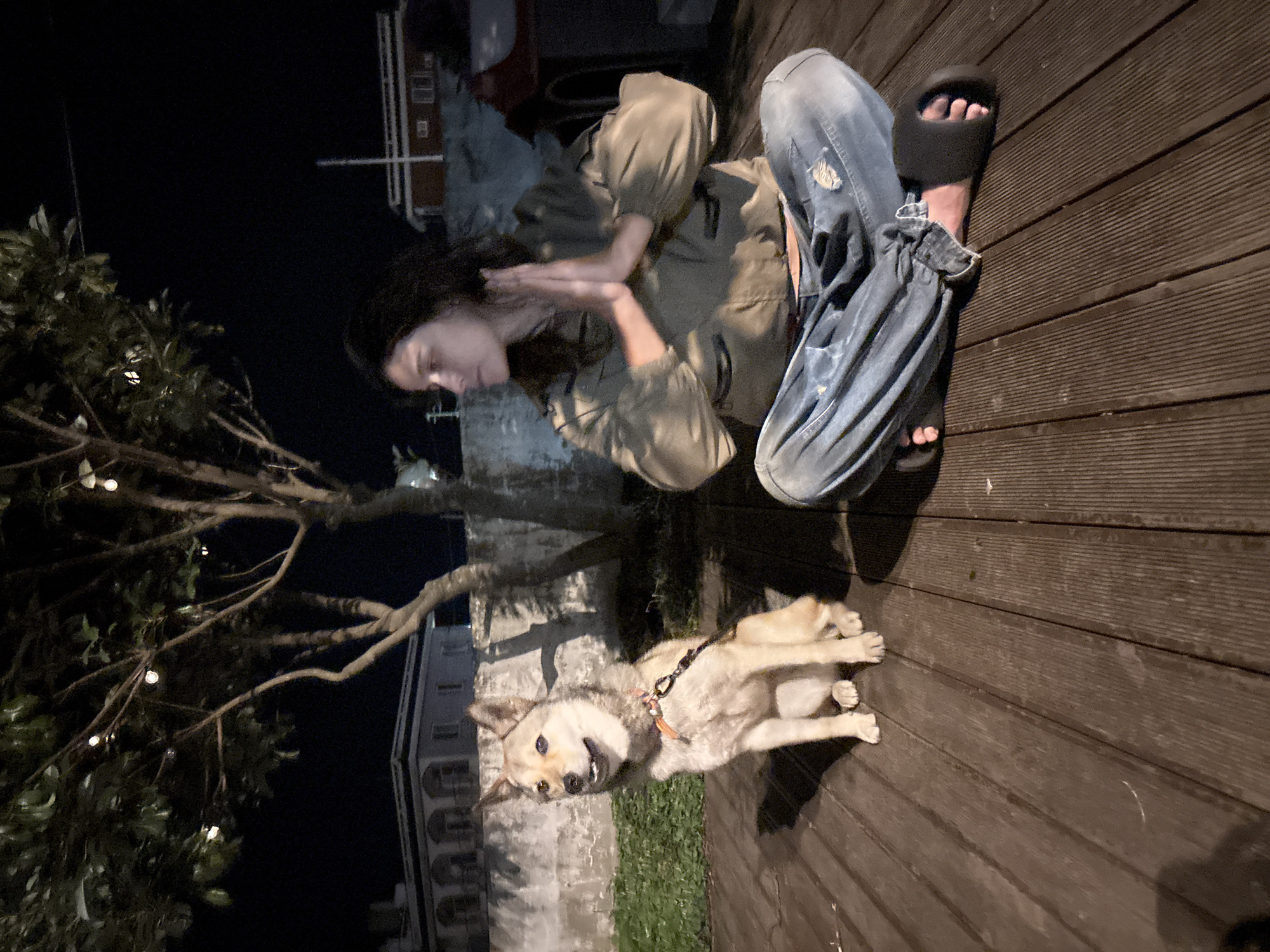 A person and a dog are on a wooden deck at night. The person is wearing a khaki jumpsuit and blue jeans, and is sitting with their legs outstretched, one foot wearing a black sandal. The dog is a light-colored medium-sized dog, lying on its side on the deck, with its head turned towards the camera. A leash is attached to the dog's collar. To the left of the deck, there is a tree with lights strung on its branches, and a building in the background. The sky is dark, suggesting it is nighttime. The overall mood appears relaxed and casual. No visible text is present in the image. The setting is likely an outdoor space, possibly a patio or garden area, in Seogwipo-si, South Korea.