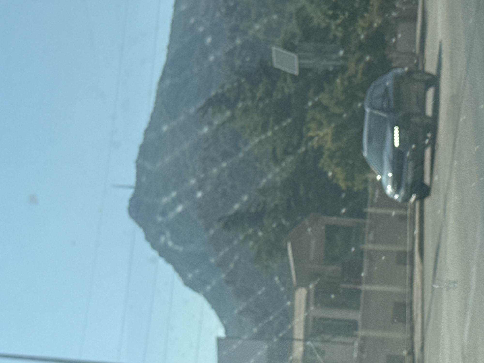 The image depicts a car driving on a road with a mountainous landscape in the background, under a clear blue sky. The mountain is covered in dense green foliage, with a visible structure on its peak. In the middle ground, there are buildings and trees. The car, which appears to be a dark-colored SUV, is in motion, with its headlights illuminated. The scene is somewhat hazy, possibly due to weather conditions or the camera lens. The overall impression is a view from a moving vehicle on a sunny day in a scenic, possibly rural, area near Sutomore, Montenegro.