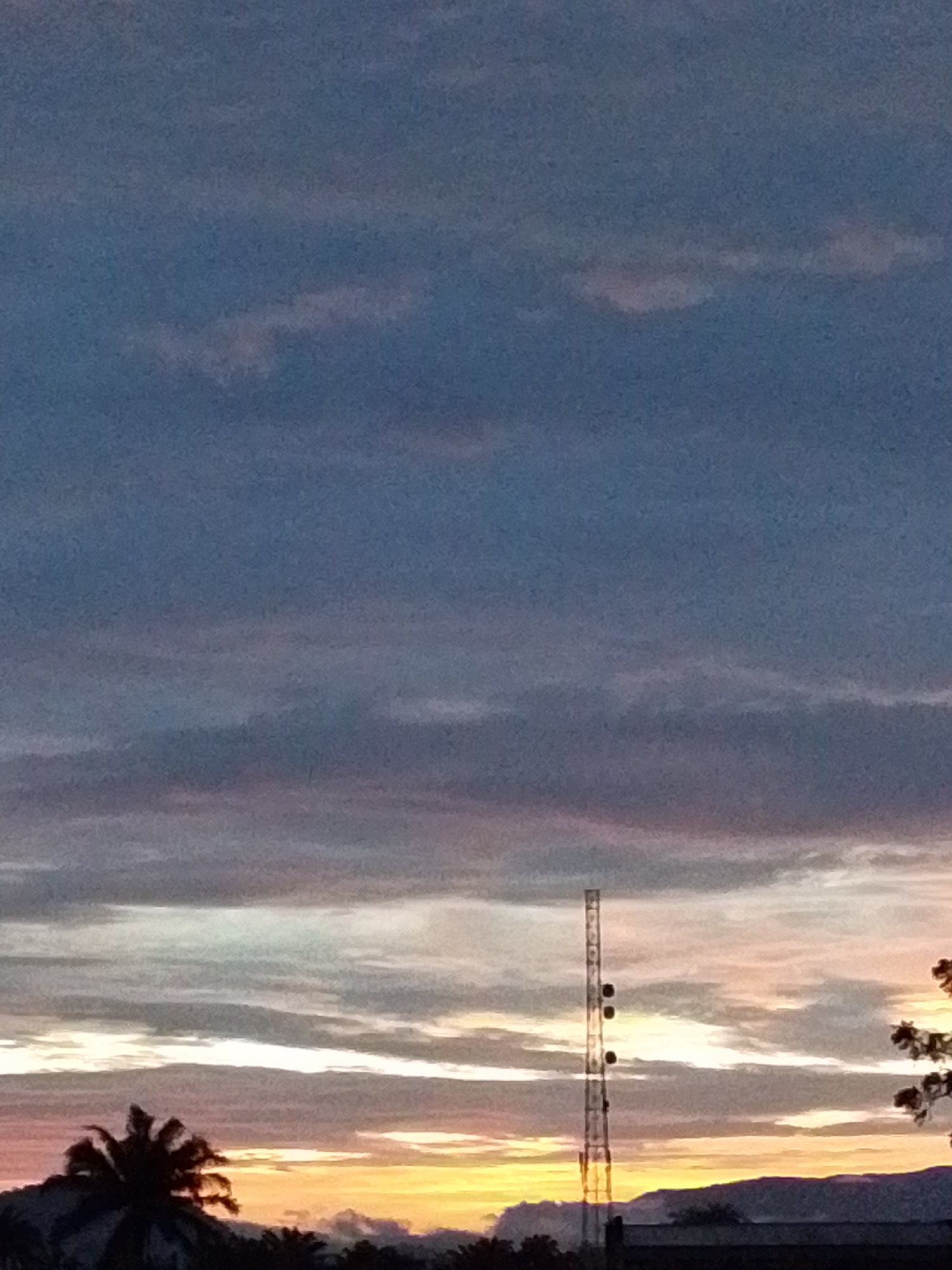 The image depicts a sunset sky over the city of Serti, Nigeria. In the foreground, silhouetted against the colorful sky, is a tall, slender telecommunications tower. The sky is filled with a gradient of colors, from deep blues at the top to hues of pink, orange, and yellow near the horizon. Clouds are scattered across the sky, catching the light of the setting sun. The lower portion of the image shows the dark silhouettes of trees, buildings, and hills. A palm tree is visible on the left side. The overall scene suggests a calm and atmospheric evening in Serti.