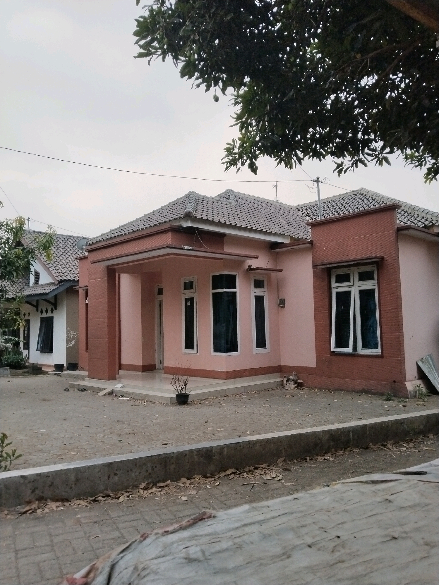 The image shows two houses side-by-side in the city of Sangkanjaya, Indonesia. The house in the foreground is painted pink with brown accents and has a gray tiled roof. It features several large windows with white frames and dark tinted glass. A small porch area with a tiled floor is visible. In front of the house, there is a paved yard with some potted plants and scattered leaves. To the left, another house is visible, painted white with dark trim and a similar gray tiled roof. This house has multiple windows and a more traditional peaked roofline. The background shows a cloudy sky and the upper branches of a leafy tree, partially obscuring the sky. Power lines are also visible against the sky. The overall scene depicts a residential area with two adjacent homes under an overcast sky.