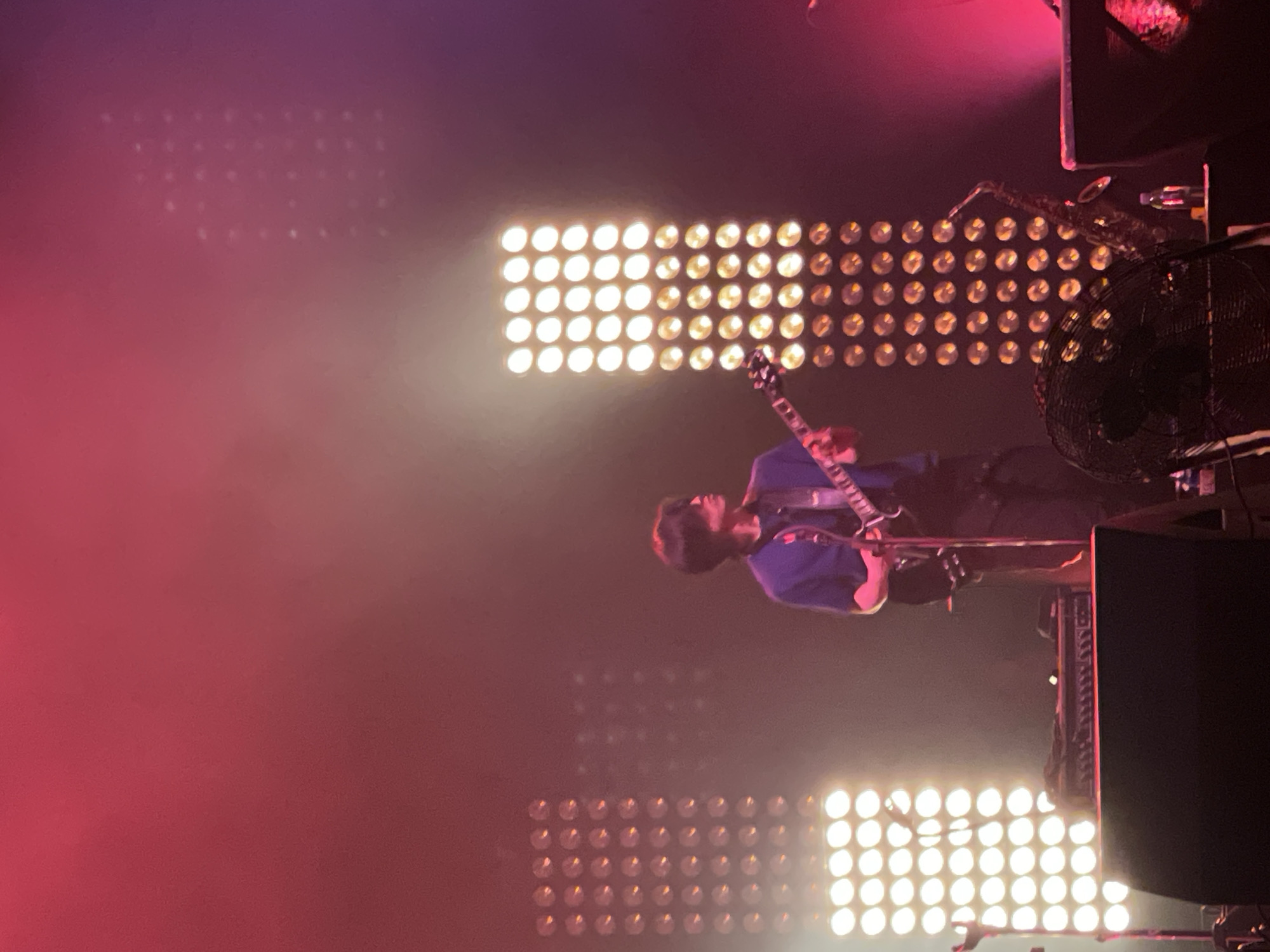 A musician is playing an electric guitar on a stage in Salembaran, Indonesia. Behind the musician are several panels of lights and a fan. A large speaker is visible to the right. The scene is lit with bright white stage lights and red lights.