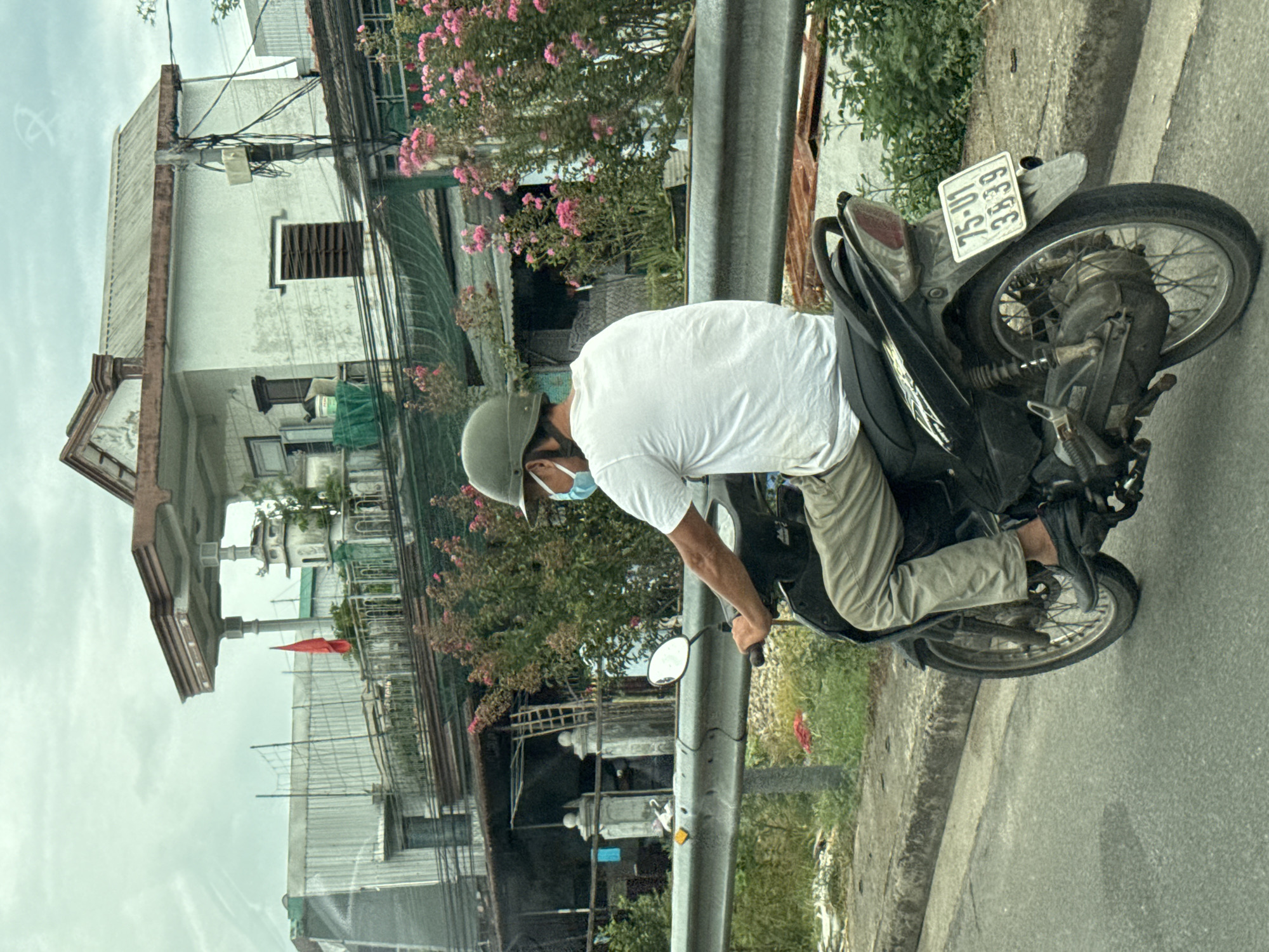 A person wearing a white t-shirt, light-colored pants, a green helmet, and a light blue face mask rides a black motorcycle. The motorcycle has a license plate that reads "75-U1 3839". The scene appears to be in Phú Xuân Ward, Vietnam, with a building featuring white walls, a corrugated metal roof, and a balcony in the background. Pink flowers are visible on bushes in front of the building. A metal railing runs along the side of the road. The sky is overcast.
