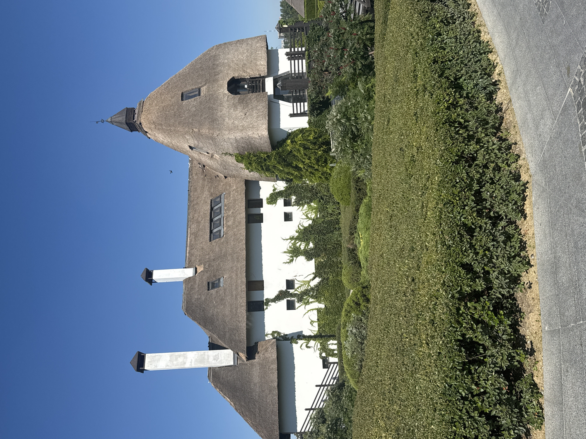The image depicts a large, white building with a thatched roof, featuring two prominent chimneys. The building is surrounded by lush green landscaping, including manicured hedges and climbing vines on the facade. A clear blue sky is visible in the background. This building is the "Chalet" restaurant, located in the Dnipro City Park in Dnipro, Ukraine.