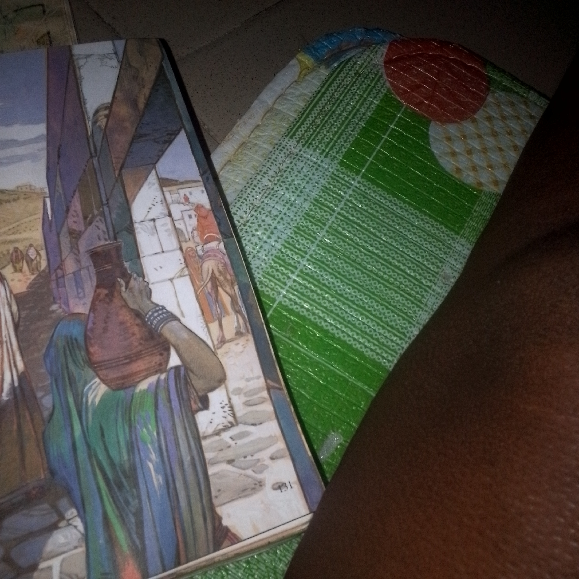 A close-up, high-angle shot captures a portion of a book featuring a painted illustration of a woman carrying a large, dark brown jug on her head, her right arm bent and supporting it. She is draped in a blue and green robe, and a beaded bracelet adorns her wrist. The background of the illustration depicts an arid, sun-baked landscape with structures and figures in the distance. To the right of the book, a folded, patterned mat with green and circular red and white sections is visible, partially obscuring a dark, textured surface that appears to be skin, likely an arm. The lighting is dim, with shadows cast across the scene.