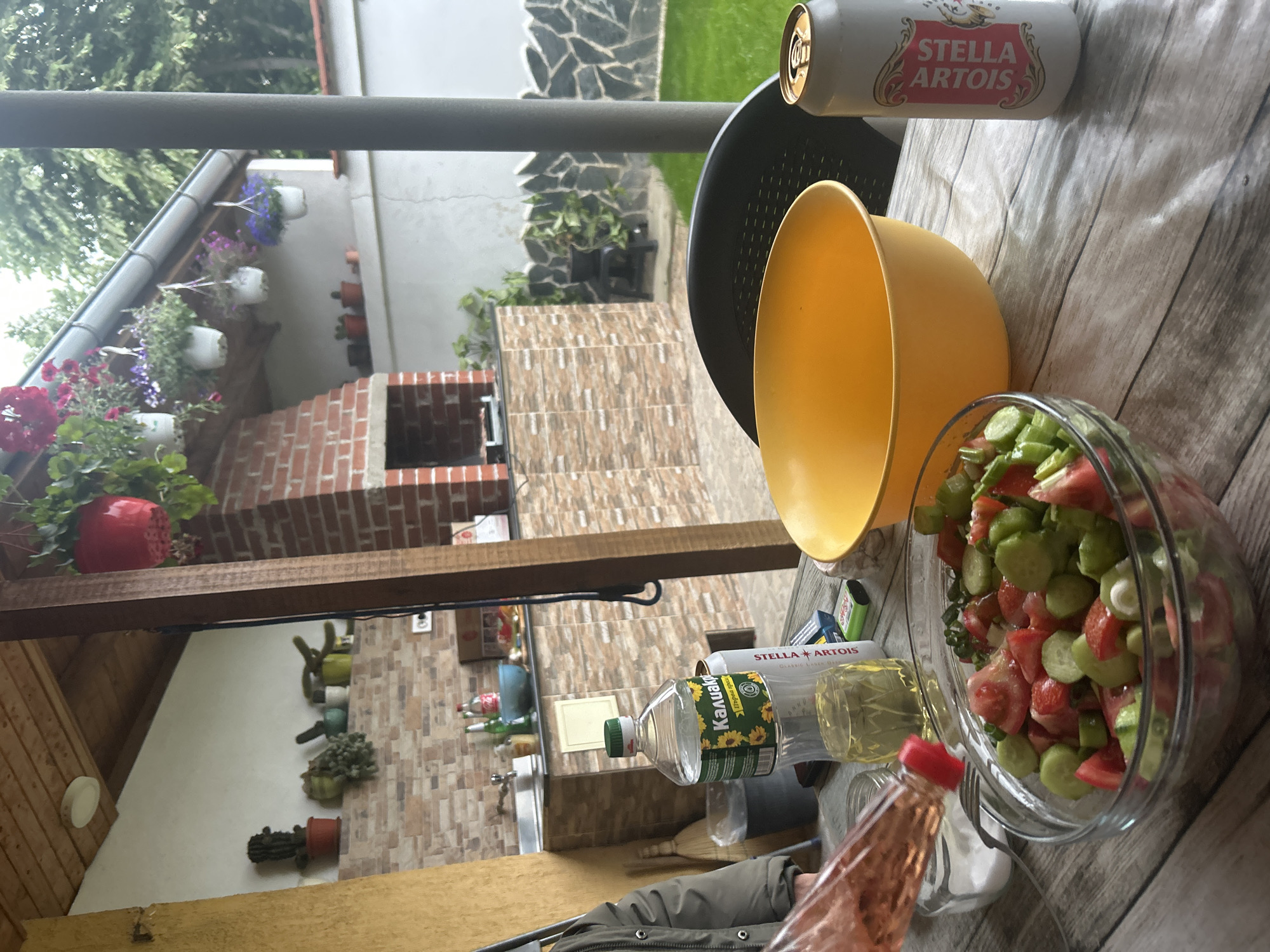The image depicts a table setting, likely outdoors, in Orizari, Bulgaria. A clear glass bowl of cucumber and tomato salad sits next to a yellow plastic bowl. A Stella Artois beer can is visible. A bottle of "Kanverk" sunflower oil and a bottle of rose vinegar, alongside salt, are present. A black plastic chair is partially visible. In the background, a brick barbecue area is decorated with flowering plants in hanging containers. A stone wall is beyond the barbecue. Inside, there is a sink. The time of day is likely daytime.