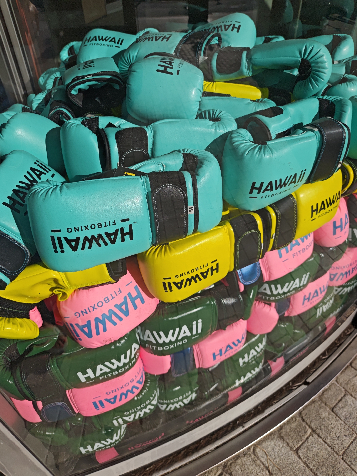 A pile of colorful boxing gloves is displayed in what appears to be a shop window. The gloves are primarily turquoise, yellow, pink, and dark green, with "HAWAII FITBOXING" printed on them in various colors. Some gloves also have a smaller black tag with "M" on them. The gloves are stacked haphazardly, creating a vibrant and textured display. The background is blurred, but suggests an indoor setting with glass reflections. The overall impression is one of a commercial display, possibly for sports equipment. The location is identified as Moscow, Russia.
