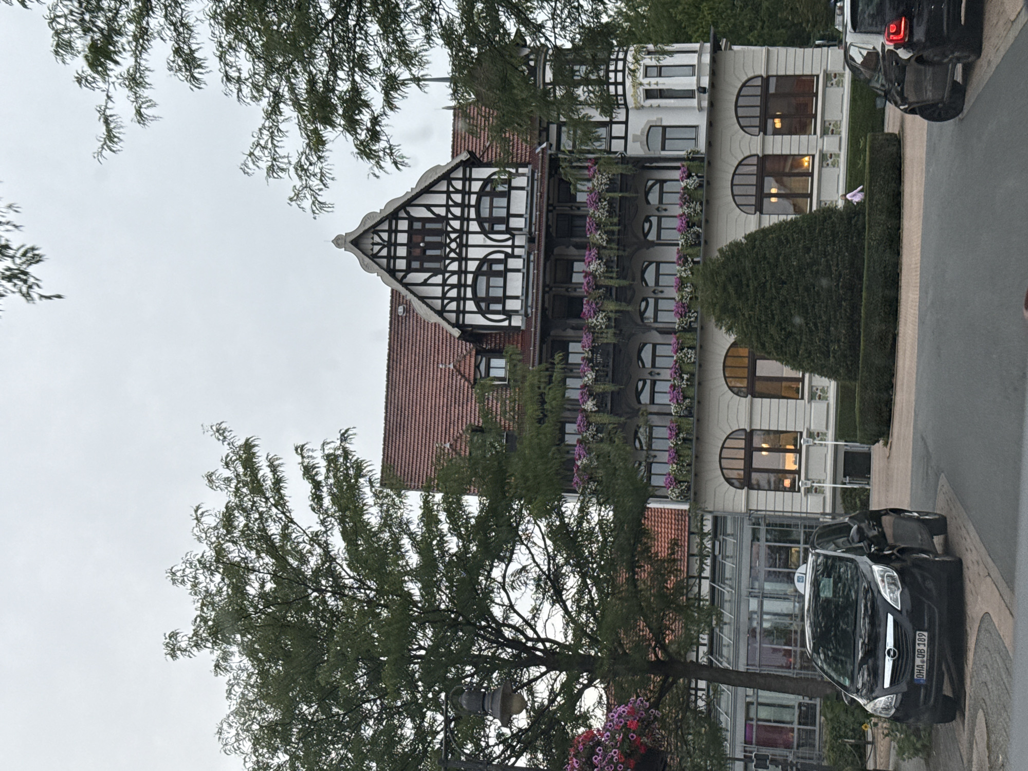 This image depicts a cloudy day in Bad Sachsa, Germany, focusing on the facade of a large, multi-story building with traditional German architecture, including timber framing and a red tile roof. The building is adorned with numerous flower boxes filled with pink and purple flowers along its balconies and window ledges. In the foreground, a black Opel Meriva is parked on a cobblestone driveway, displaying a license plate that reads "OHA QB 189". To the right of the Opel, another black vehicle, partially visible, is parked. A person in a light-colored outfit is seen walking in the middle ground in front of a large, cone-shaped evergreen bush and manicured hedges. Several trees with green foliage frame the scene, with one prominent street lamp visible on the left side of the image, also adorned with flowers. The overall environment suggests a hotel or a similar establishment in a well-maintained, possibly park-like setting within the town of Bad Sachsa.