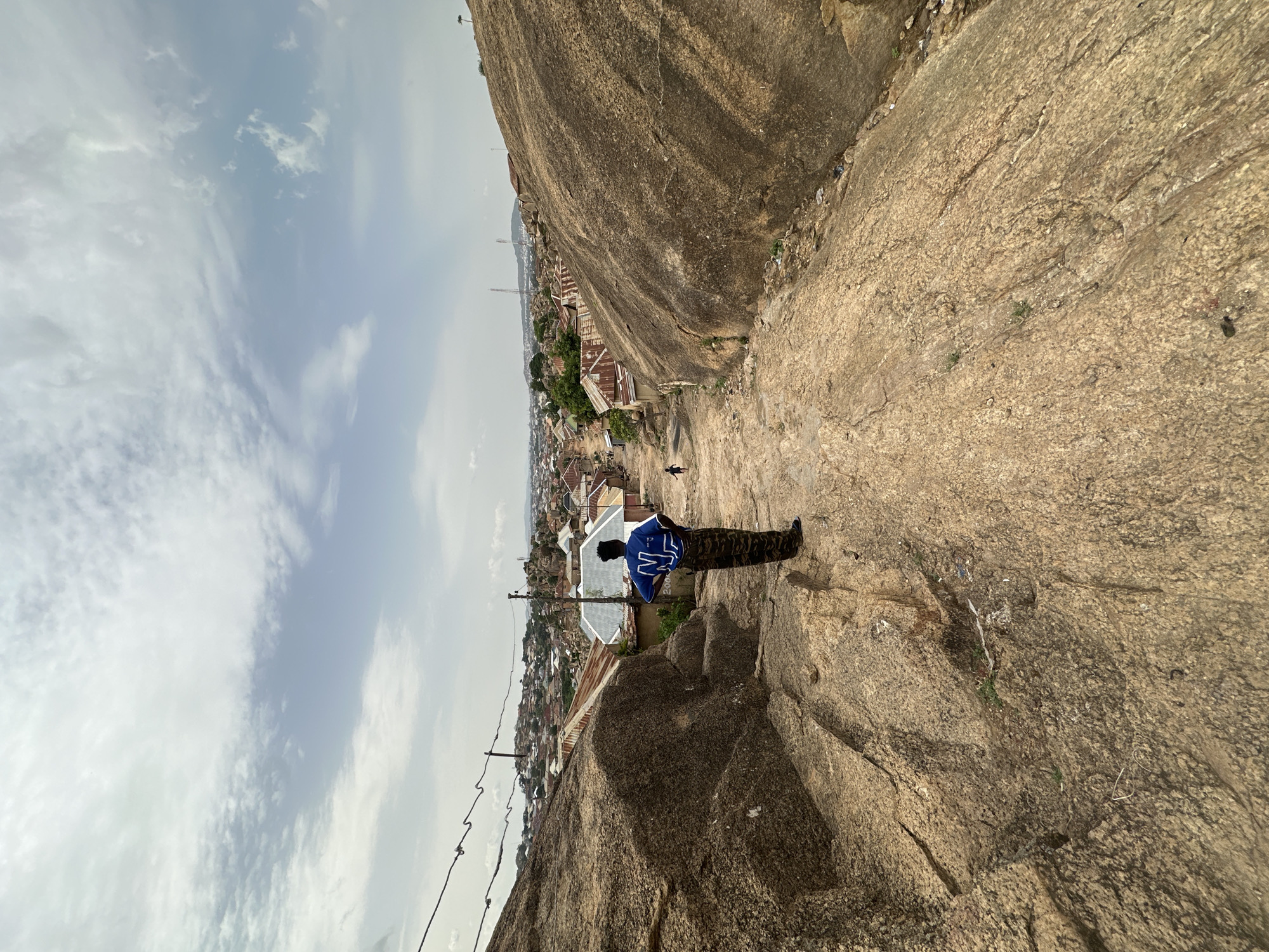 An eye-level photograph shows a person standing on a light brown rocky cliff, appearing to look out over the city of Jos, Nigeria. The person is facing away from the camera and wearing camouflage pants and a blue shirt with white lettering on the back. The city is nestled in a valley, featuring buildings of varying sizes and construction. Power lines run down the cliff in the foreground. The sky overhead is partly cloudy. The photograph is rotated on its side.