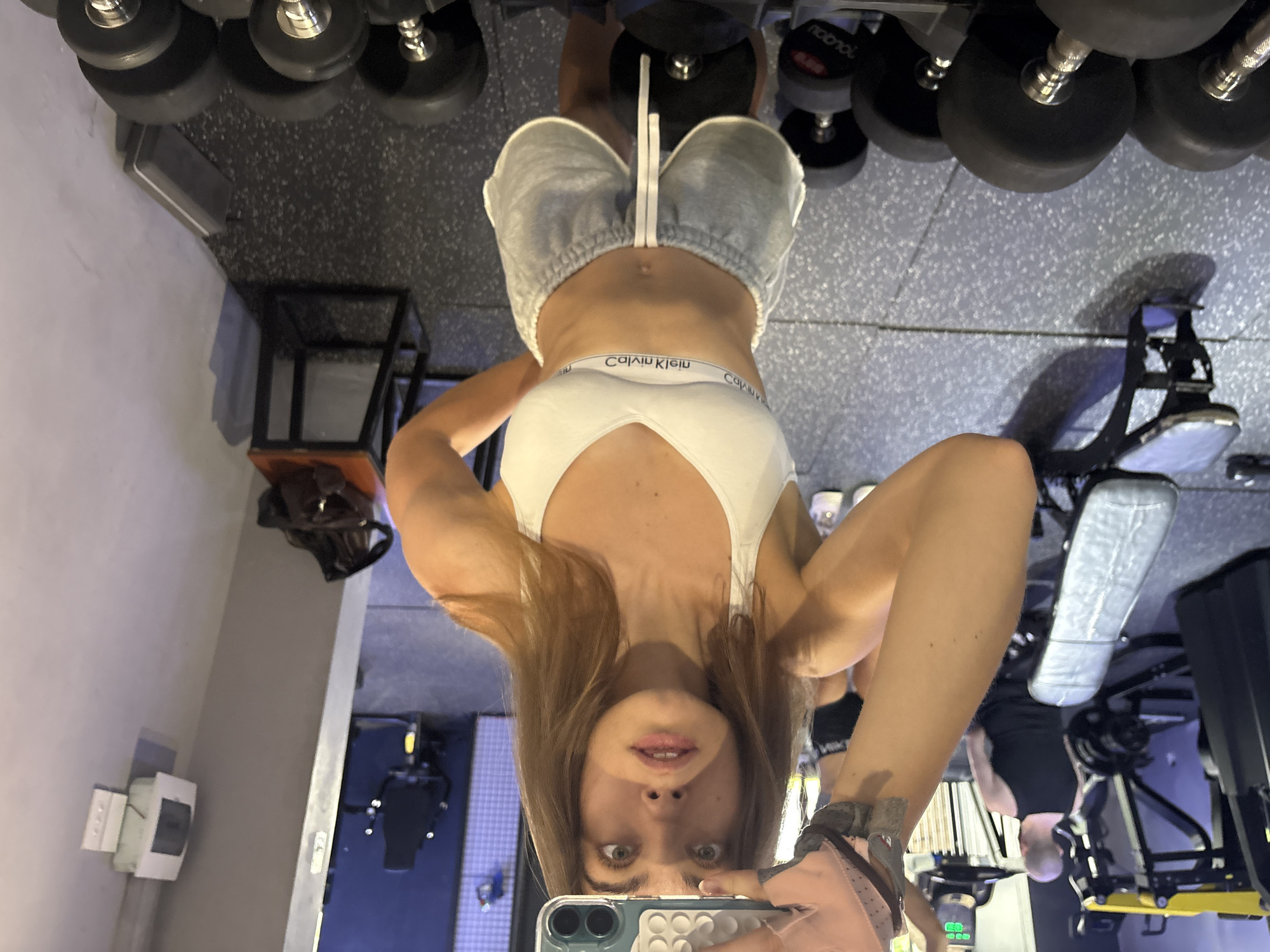 A woman in a white sports bra and gray shorts is taking a selfie in a gym. Dumbbells are above her head. Gym equipment can be seen in the background. The location is likely a gym, but the specific location in Đà Nẵng, Vietnam is not discernible from the image.
