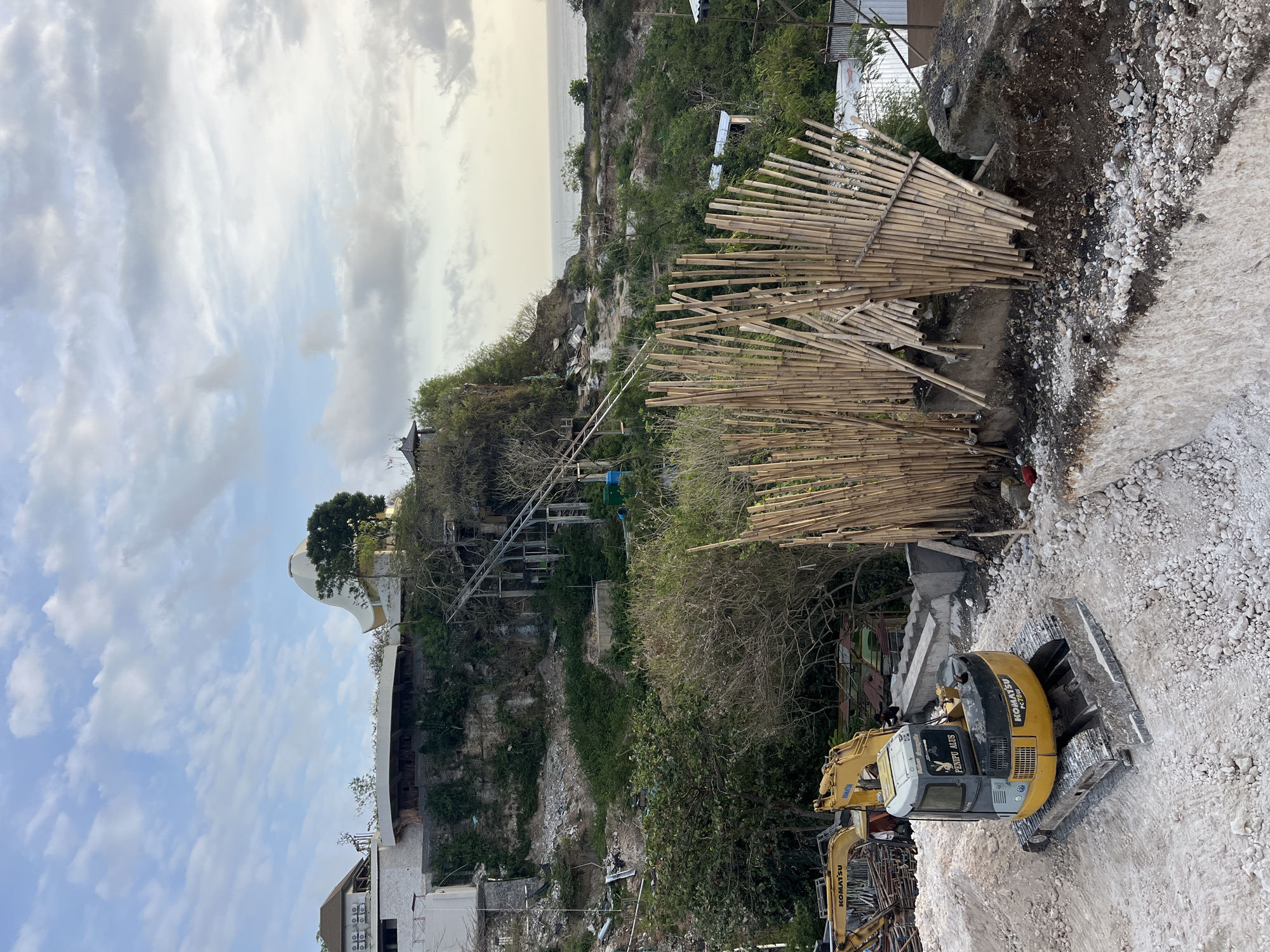 The image captures a construction site in Kuta Selatan, Indonesia, with a view of the ocean in the background. A yellow Komatsu excavator is parked on a mound of light-colored gravel and debris in the foreground. Behind the excavator, a partially constructed building with a distinctive white, curved roofline is visible on a hillside, surrounded by lush greenery. Several other structures and scaffolding are also present on the elevated terrain. In front of the hillside, a rustic fence made of bamboo poles is erected. The sky is partly cloudy, with the sea stretching out to the horizon under a soft, diffused light.
