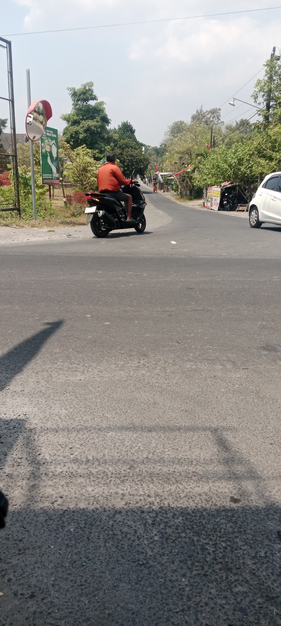 A person wearing an orange shirt and dark shorts is riding a black scooter on a paved road. The scooter has a white license plate. The road appears to be in a rural or suburban area, with trees and vegetation lining the sides. In the background, there are buildings, including a small shop with motorcycles parked outside. A white car is also visible on the right side of the road. A red convex mirror is mounted on a pole on the left side of the road, reflecting part of the scene. A sign with text and an image is also visible near the mirror. The sky is partly cloudy. The image is taken from a low angle, with the shadow of the camera or person taking the photo visible on the road in the foreground. The location is specified as Maguwoharjo, Indonesia.