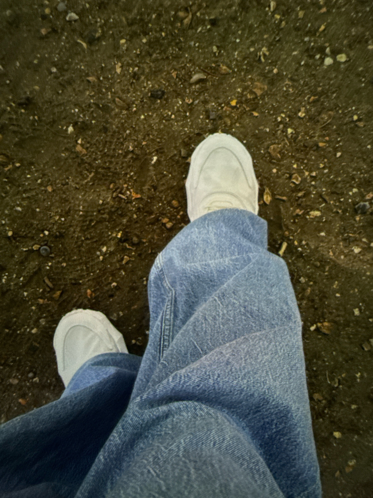 A close-up, overhead view captures a person's legs and feet wearing light-colored sneakers and blue denim jeans, standing on a surface of dark soil mixed with small pebbles and debris. The setting appears to be an outdoor urban environment, possibly a park or a walkway in Basildon, United Kingdom. The lighting suggests it is either dusk or an overcast day, as there are no strong shadows. The composition focuses on the textures of the clothing and the ground, with the feet positioned towards the lower center of the frame.