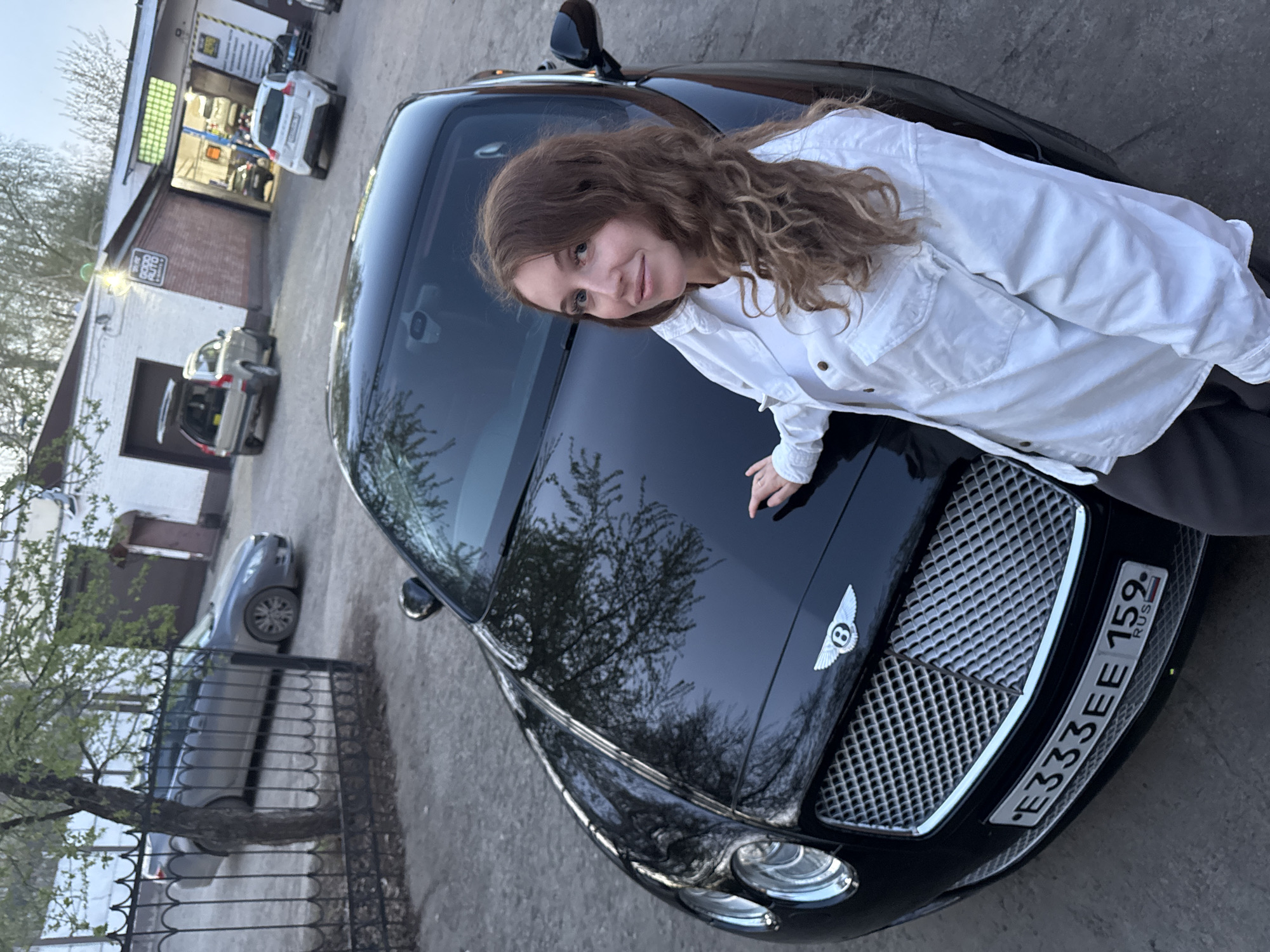 A woman with long, curly brown hair is leaning on the hood of a black Bentley car. She is wearing a white button-down shirt and dark pants. The car has a Russian license plate that reads "E333EE 159."
The scene is set in Chaikovsky, Russia, as indicated by the license plate code (159). In the background, there is a brick building with a sign that reads "Робо Авто". There are other cars parked in front of the building. A metal fence and a tree are visible on the left side of the image.