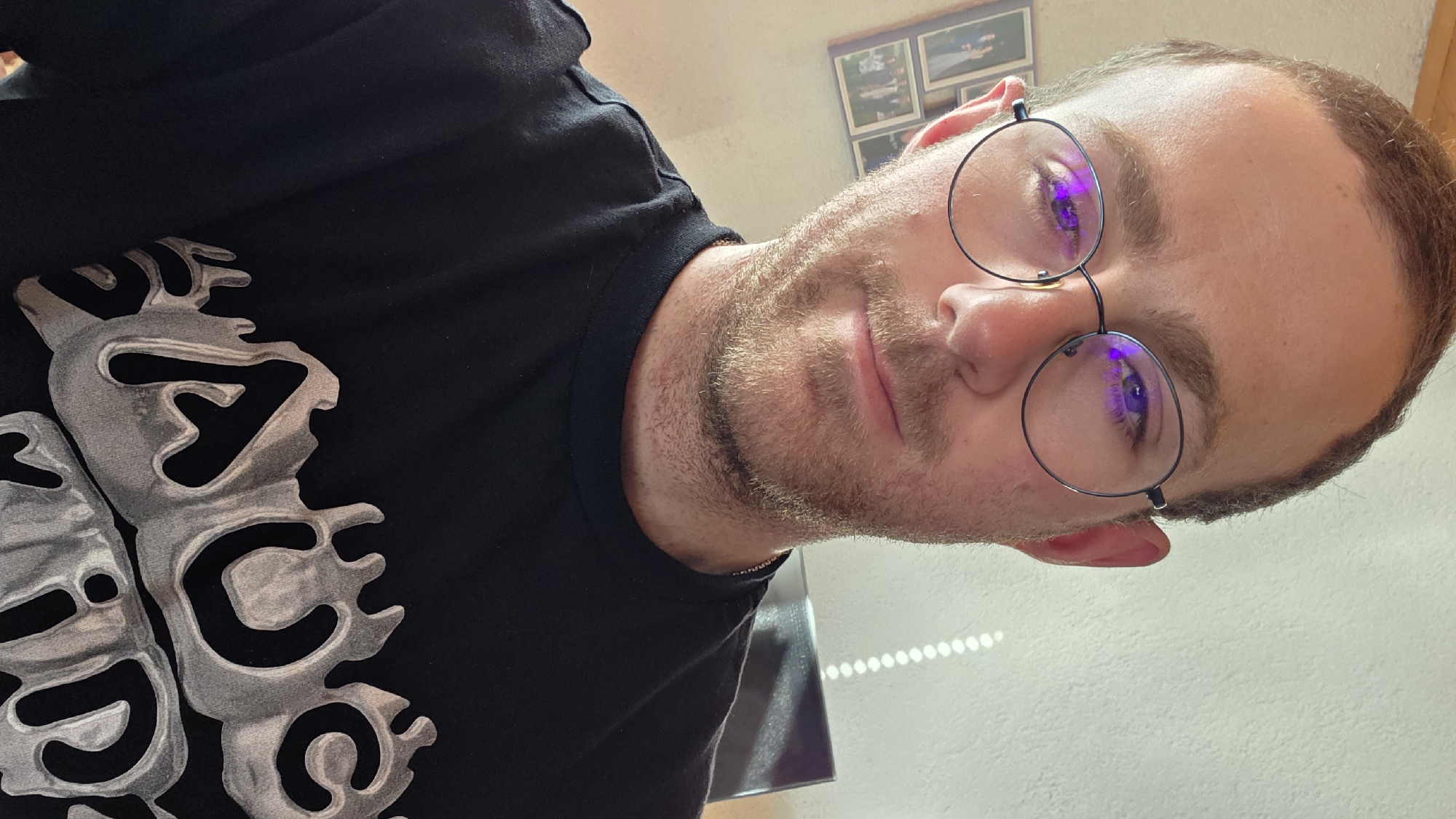 A selfie shows a man with short brown hair, a light beard, and round glasses. He is wearing a black t-shirt with a white and gray design. The background appears to be an indoor setting with a white wall and some photos in frames hanging on the wall. A light source creates a linear pattern of light on the wall. The man's reflection is visible in his glasses.