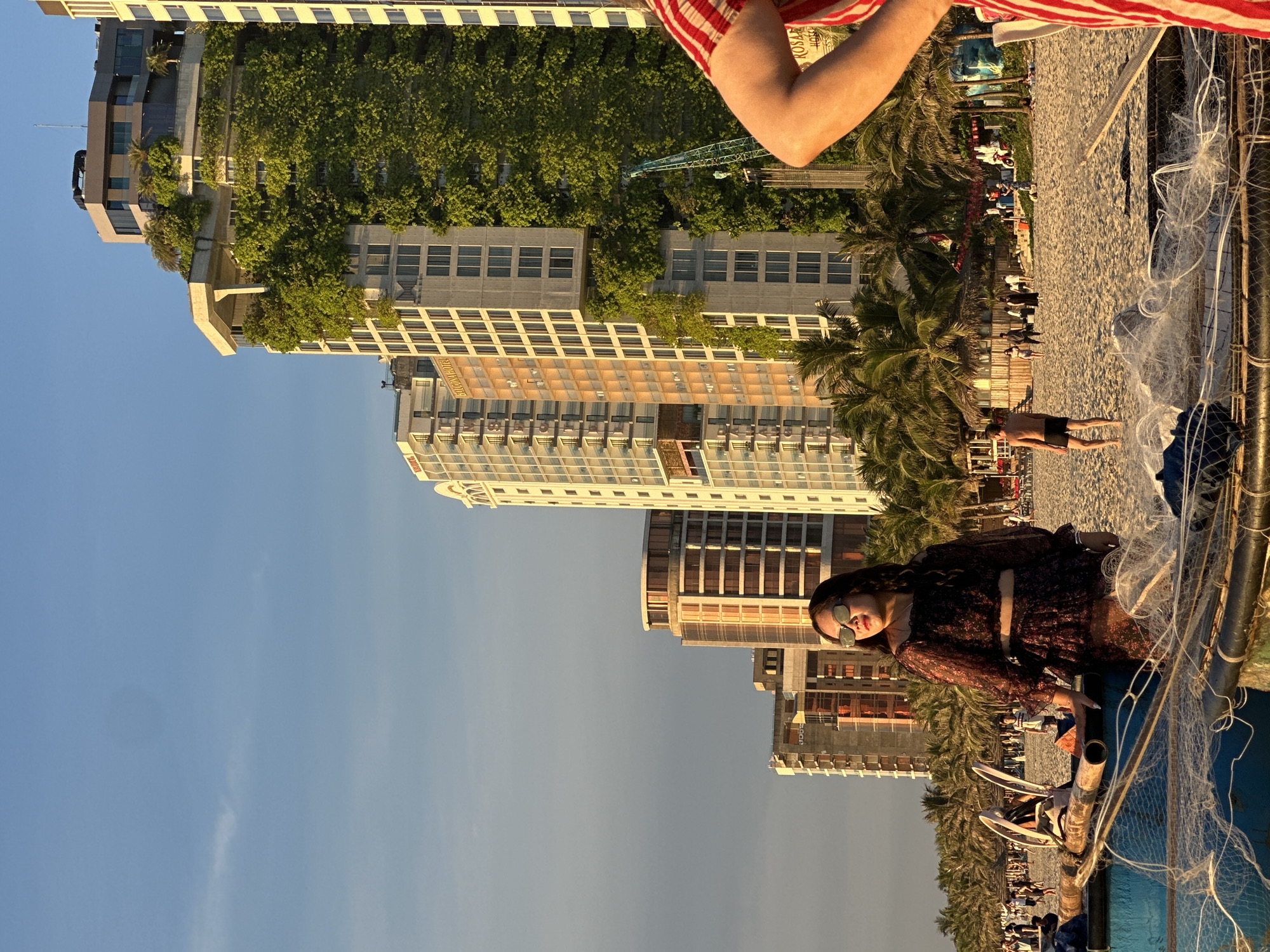 The image depicts a beach scene in Đà Nẵng, Vietnam, with a prominent row of modern high-rise buildings in the background. The buildings are architecturally diverse, with one exceptionally notable structure covered in lush green vegetation. In the foreground, a woman in a dark floral dress and sunglasses stands near a traditional blue fishing boat laden with fishing nets. Several other people are visible on the sandy beach, some walking and others relaxing. Palm trees line the beach, interspersed with various beach umbrellas and small structures. The sky is a clear, soft blue with a hint of clouds, and the golden light suggests it is either early morning or late afternoon. A portion of another person's arm, wearing a red and white striped shirt, is visible in the far right of the frame.