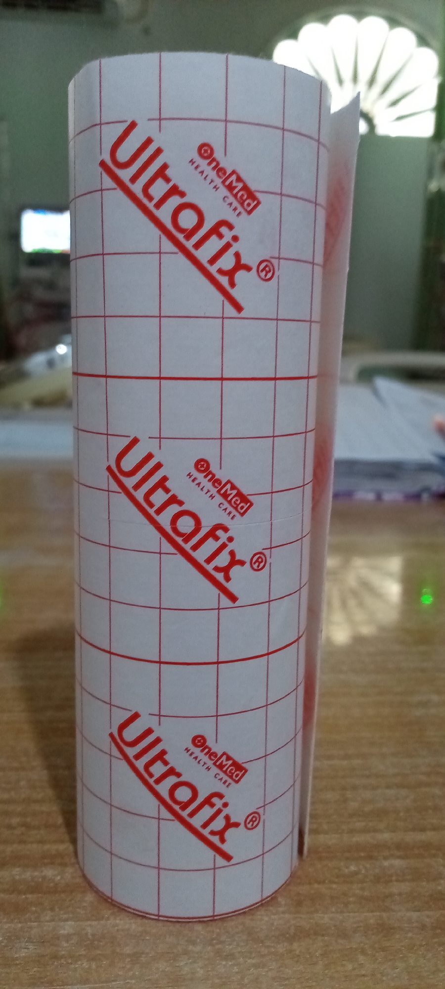 A roll of transparent adhesive medical tape stands on a wooden surface. The tape is printed with the words "Ultrafix" and "One Med Health Care" in red. The tape has a grid pattern, also in red. The background is blurred, but a television screen is visible to the left, and a semi-circular decorative light fixture is visible in the upper part of the image. The picture was taken in an indoor environment. The geographical location of Lhokseumawe, Indonesia, is not relevant to the image.