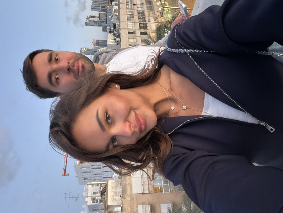 The image depicts a selfie of two individuals, a female in the foreground and a male behind her, positioned on a rooftop or elevated balcony overlooking an urban landscape in Tel Aviv, Israel, during late afternoon or early evening conditions.
The female subject, centered and facing the camera with a slight smile, has long brown hair and is wearing a dark blue zippered jacket over a white top. She has two silver necklaces, one featuring a small clover pendant and another with a square pendant on a Y-shaped chain. A single diamond stud earring is visible in her right ear. The male subject, partially visible behind her right shoulder, has short dark hair and a neutral expression, looking directly at the camera. He wears a light-colored t-shirt under a grey hooded jacket. A silver-colored smartphone is partially visible in his right hand on the right side of the frame.
The background details an urban environment. Immediately behind the subjects are multiple multi-story buildings of older construction, characterized by flat roofs, visible air conditioning units, and windows, some with external roller shutters. To the left, a red construction crane is visible above a white building undergoing construction. In the distant background, a cluster of modern high-rise buildings forms a city skyline. The sky is a light blue, transitioning to a softer, more muted hue near the horizon, indicative of twilight. A palm tree frond is partially visible in the lower-left corner of the frame.