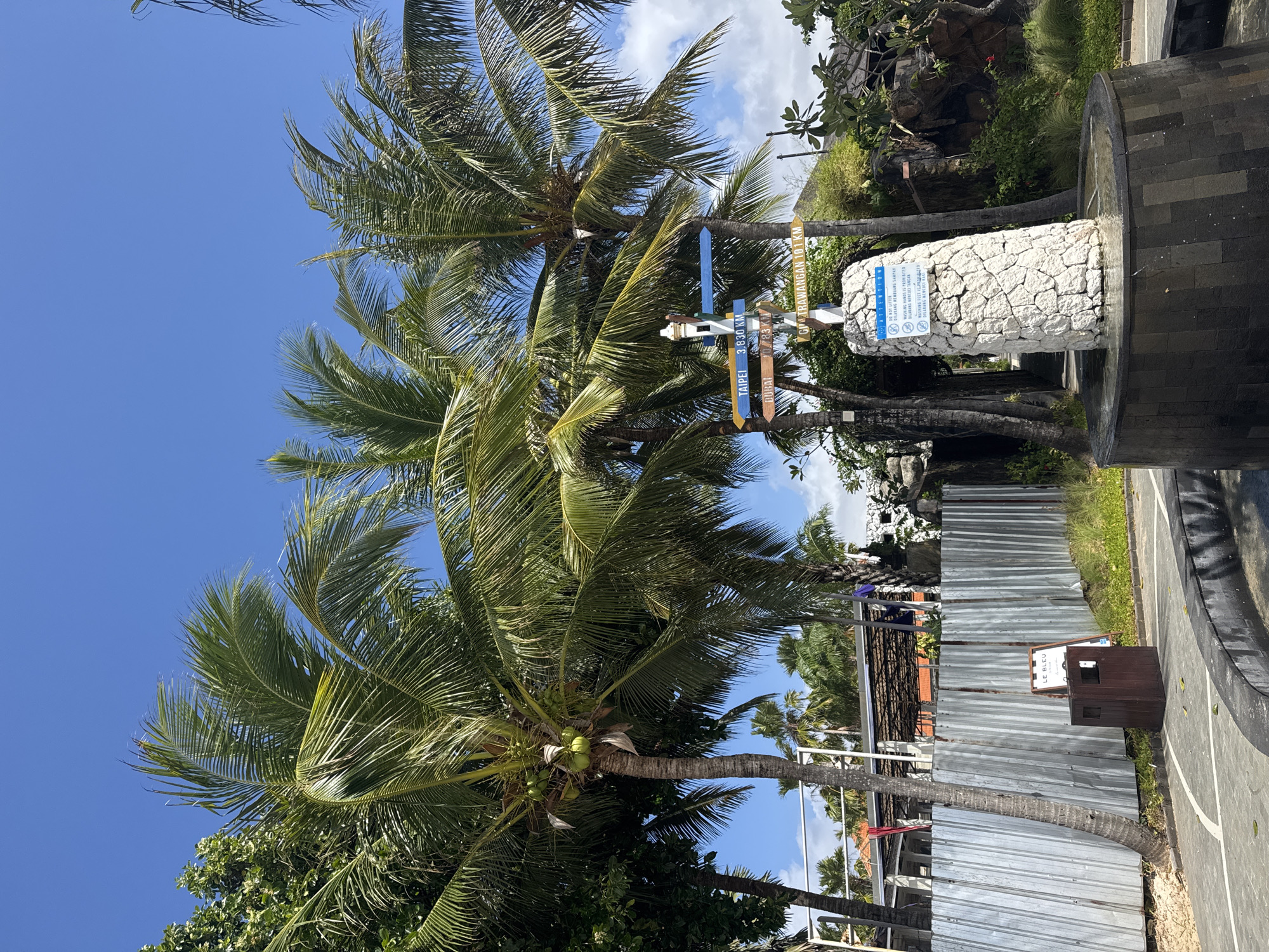A directional signpost stands in front of a fountain at a resort. The signpost has multiple signs indicating distances to various locations, including Taipei, Dubai, and Gili Trawangan. Palm trees surround the area, and a metal fence is visible in the background, along with a wooden trash receptacle and a sign that reads "LE BLEU." The sky is clear and blue, with some white clouds. The scene appears to be a tropical or subtropical location.