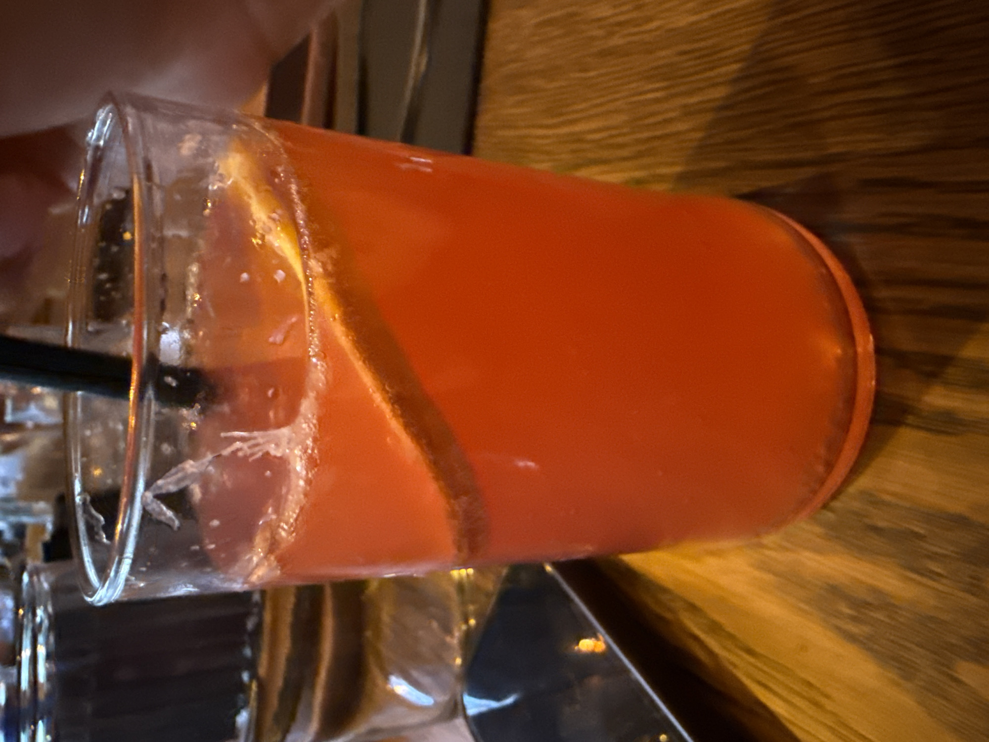 An orange beverage is served in a clear glass with a black straw and a slice of orange. The glass sits on a wooden table. In the background, another glass is visible, as well as a dark rectangular object, possibly a phone. The scene is dimly lit, with the warm glow of the table suggesting an indoor setting. The image does not provide enough information to determine the specific location in Sofia, Bulgaria.