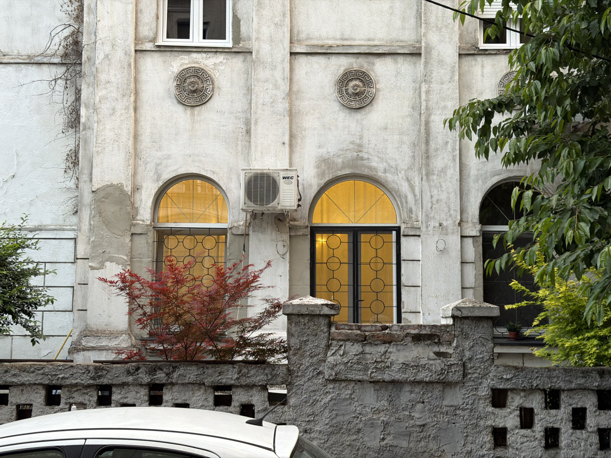 Exterior view of an aged, pale-colored building with architectural details like decorative round reliefs and arched windows. An air conditioning unit is mounted on the facade between two of the windows. A low, weathered concrete wall runs across the foreground. A red-leaved shrub grows in front of one window and a tree with green leaves is partially visible on the right. The roof of a white car is visible in the lower part of the frame. The location is Vracar Urban Municipality, Serbia.
